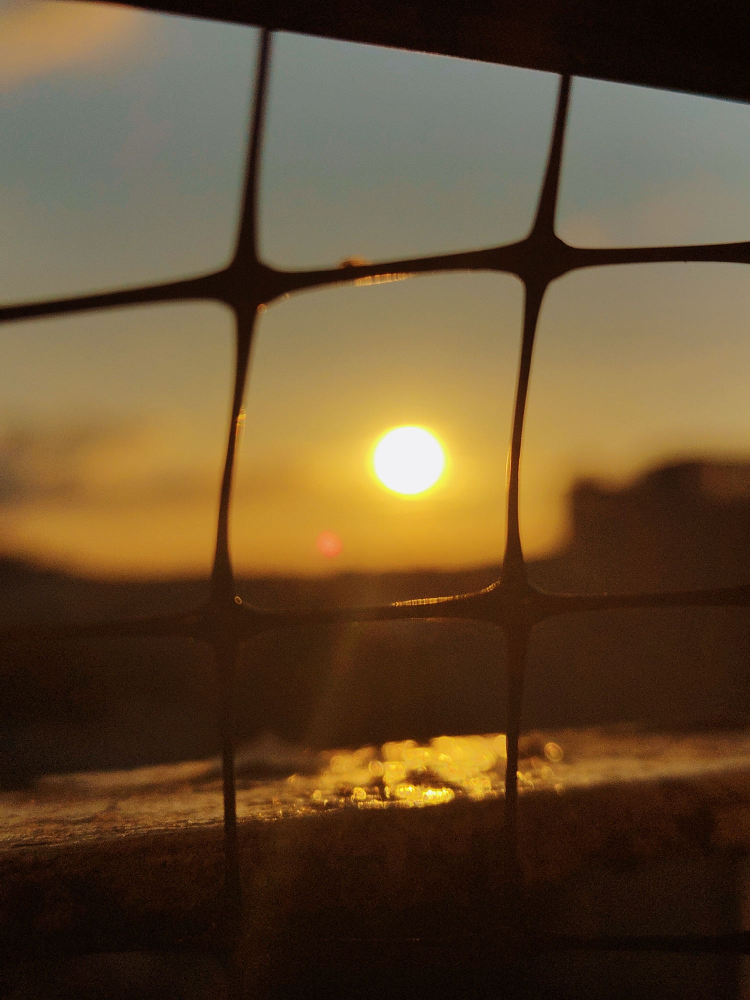 The image depicts a sunset viewed through a window with a dark, grid-like obstruction. The sun, a bright, warm orb, is positioned in the center of the frame, casting a golden glow across the sky and the reflective surface below. The foreground is dominated by the blurry, dark bars of the grid, creating a sense of looking through a barrier. The lower portion of the image shows a dark, textured surface that reflects the sunset's light, creating shimmering patterns. In the distance, indistinct shapes suggest a cityscape or landscape, silhouetted against the warm hues of the sky. The overall impression is one of peaceful contemplation, with the grid acting as a frame for the natural beauty of the sunset. The location is identified as Nowy Dwór Mazowiecki, Poland.