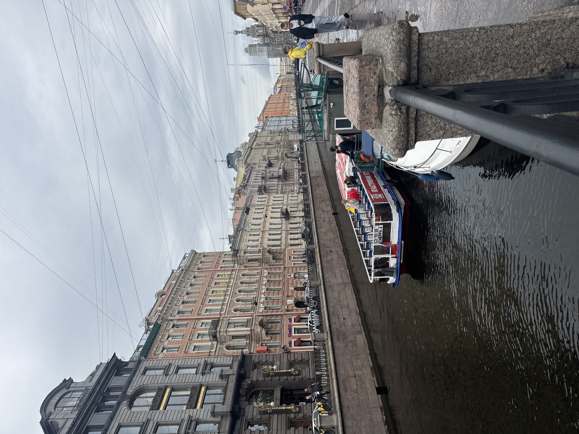 This is a photograph of the Griboyedov Canal in Saint Petersburg, Russia, on a cloudy day. The canal is lined with historic multi-story buildings in various shades of pink, beige, and gray, featuring ornate architectural details. Several boats are docked or navigating the canal. In the foreground, a granite embankment with a black metal railing overlooks the water. A white motorboat is tied up next to a larger red and white tour boat filled with passengers. On the embankment, a young man in a black jacket and jeans stands looking out, while another young man in a bright yellow jacket stands beside him. Further down the embankment, several other individuals are walking. In the distance, the iconic Church of the Savior on Spilled Blood with its green domes is visible. Overhead, a complex network of telephone or electrical wires crisscrosses the sky. The wet cobblestone sidewalk suggests recent rain.