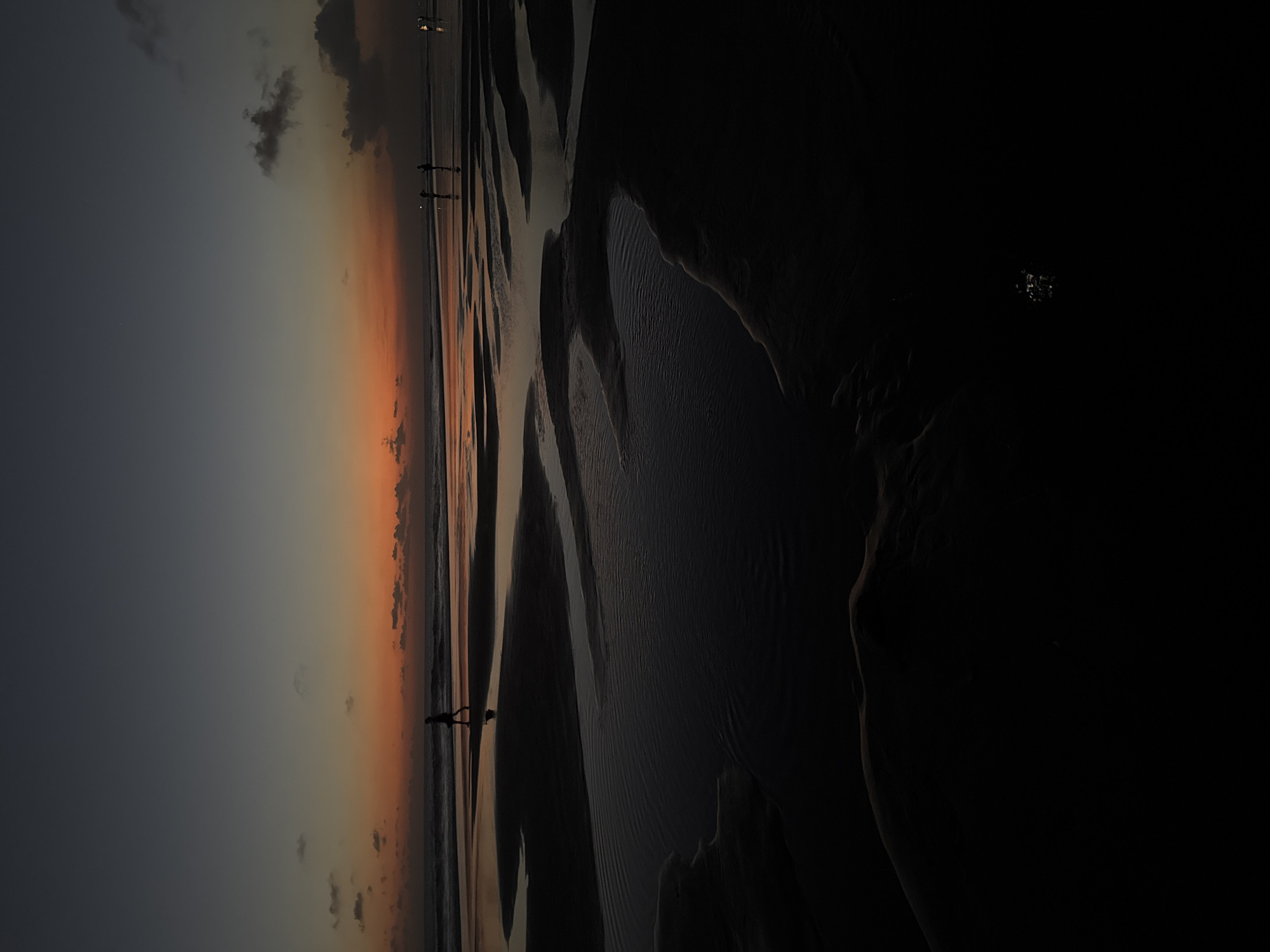 The image depicts a twilight scene at Kuta Beach, Bali, Indonesia. The sky exhibits a gradient of dark gray at the top transitioning to vibrant orange and yellow hues closer to the horizon, indicating sunset or sunrise. Several people are visible as silhouettes on the wet sand near the water's edge. One person walks alone on the left side of the frame, their figure clearly defined against the brighter sky and reflecting on the wet sand. Further in the distance, two other figures stand together, and on the far right, a small group of people are illuminated by lights, suggesting they are near an establishment. The foreground is dominated by dark, wet sand with intricate patterns formed by receding water, creating pools and channels that reflect the dim light. The overall impression is one of a serene and tranquil moment on the beach as daylight fades.