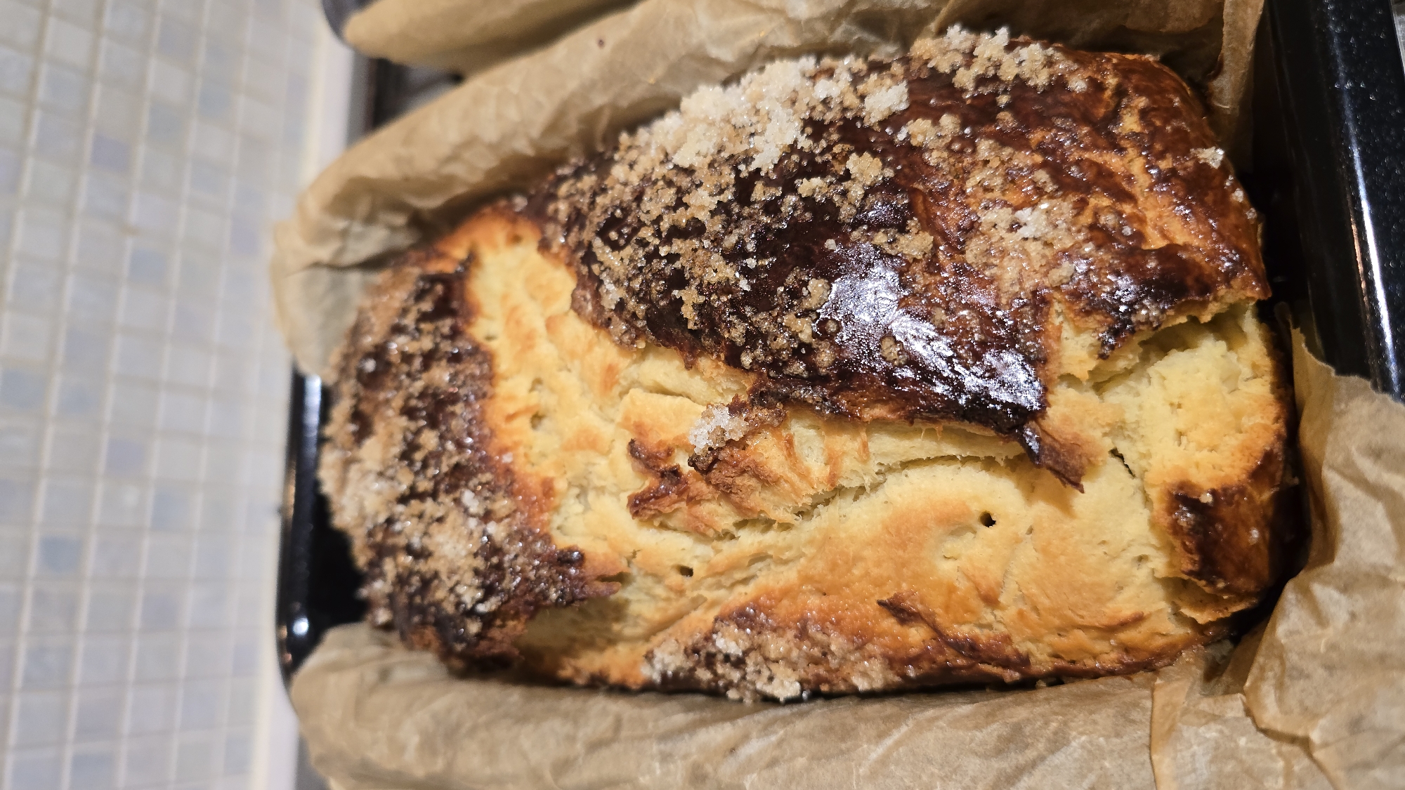 A close-up shot presents a freshly baked pastry, likely a sweet bread or cake, sitting in a dark baking pan lined with brown parchment paper. The pastry has a golden-brown crust with a dark glaze on top, sprinkled with coarse sugar crystals. The interior of the pastry reveals a soft, yellow, doughy texture. The precise location and city are not discernible from the image.