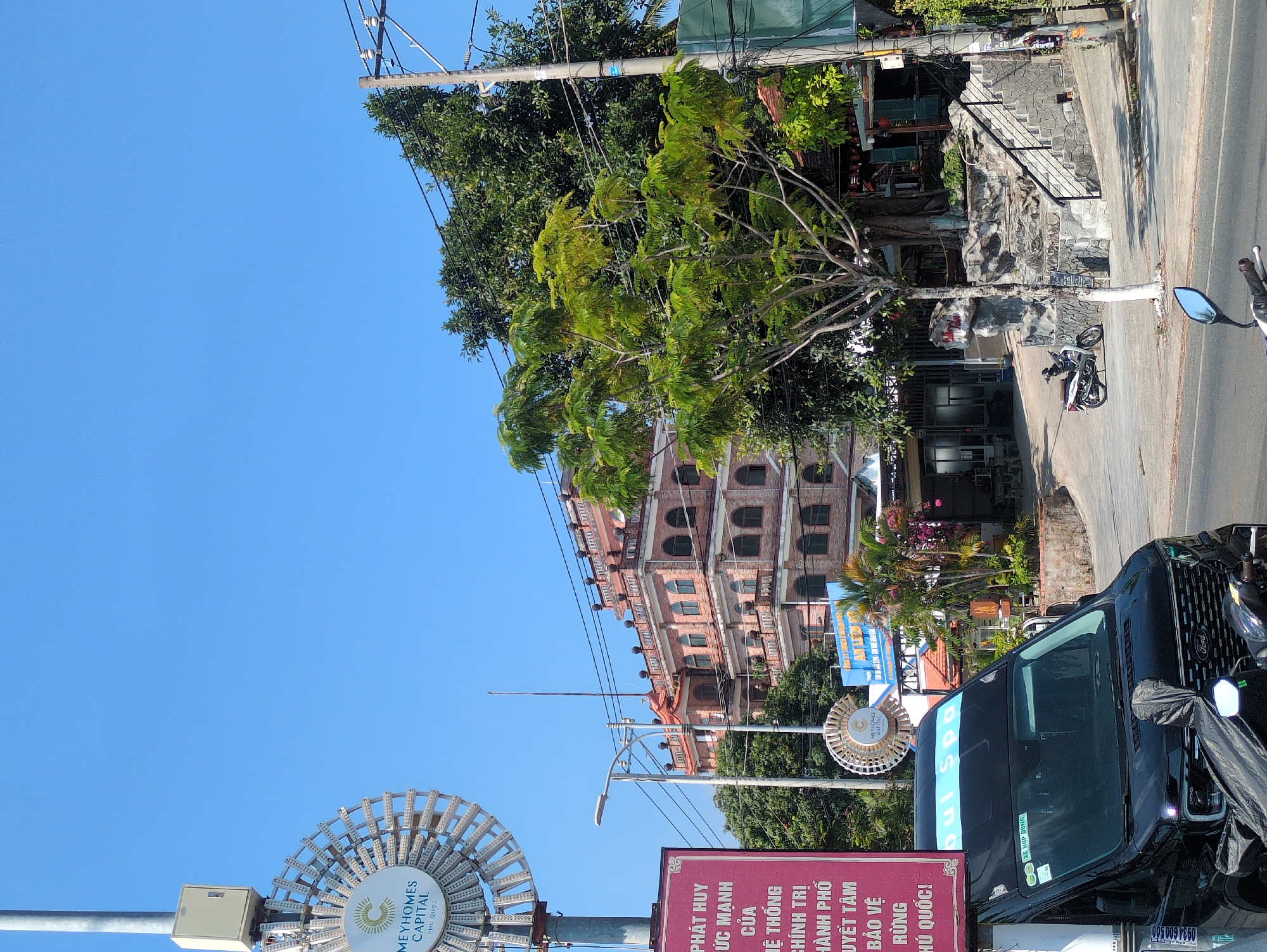 The image shows a street scene in Phú Quốc, Vietnam, with a view of a multi-story brick building and various signage.
In the foreground, on the left, is a Meyhomes Capital Phu Quoc advertisement sign with a white decorative element resembling a stylized sun. Beside this, there is another smaller sign in red Vietnamese text. A black Ford van is parked on the right side of the street, with a blue banner displaying "Dui Spa." The vehicle obscures the lower portion of the building behind it.
Across the street, the brick building features multiple arched windows and decorative elements. A tree with green leaves partly obscures the building. A parked motorbike is visible in the middle ground.
The sky is clear and blue. Overhead power lines are visible.