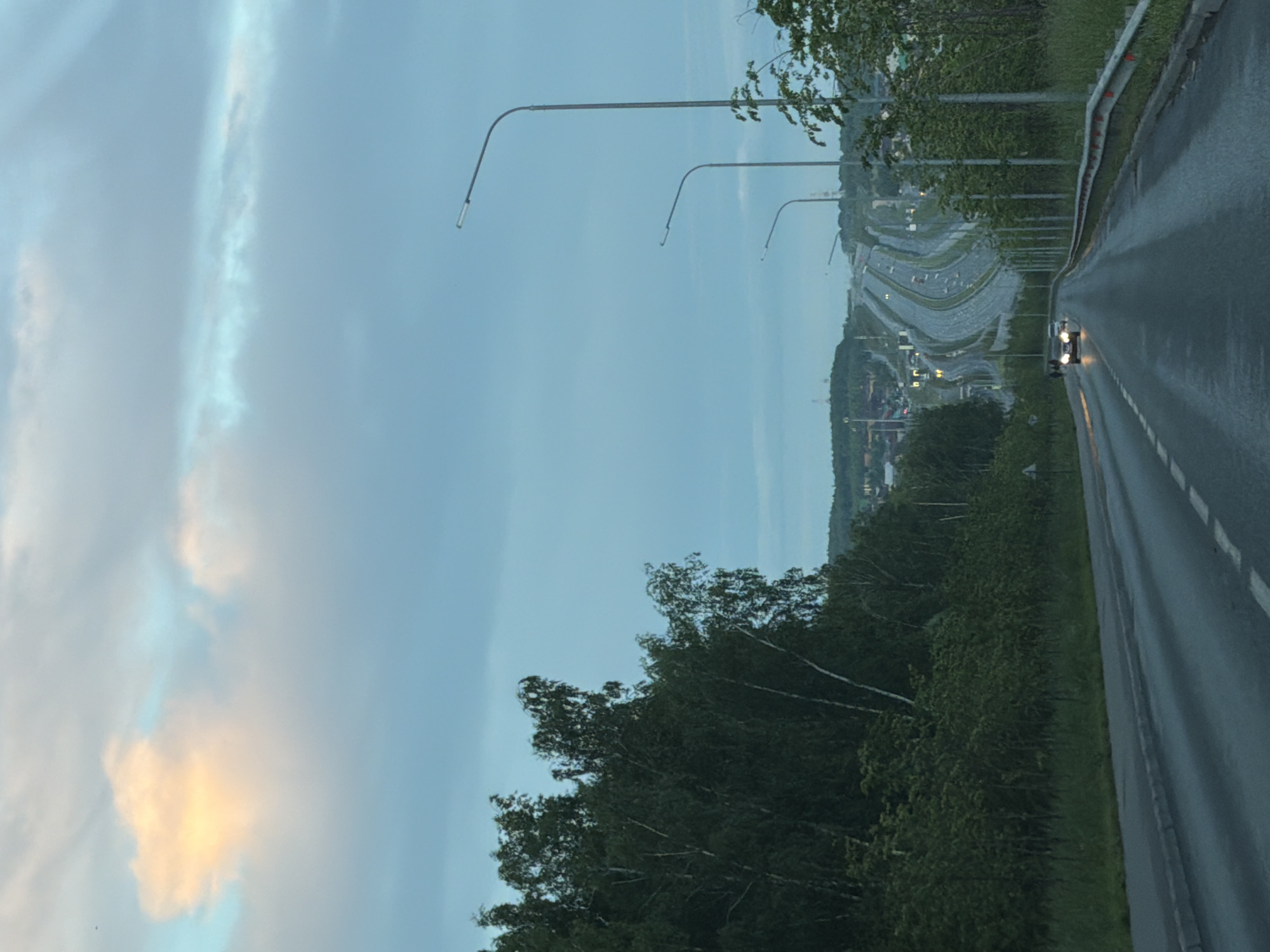 In the city of Unknown, Unknown, a highway curves uphill under a hazy, overcast sky. A single, bright orange cloud is visible in the upper left corner. Tall streetlights line the right side of the road, and a dense line of dark green trees and birch trees borders the left. The asphalt road is wet and reflects the dim light, with white dashed lines marking the lanes. Two cars are visible on the highway, one in the foreground with headlights on, and another further up the road. In the distance, houses and buildings are scattered across rolling hills. The scene appears to be at dusk or during a period of low light.