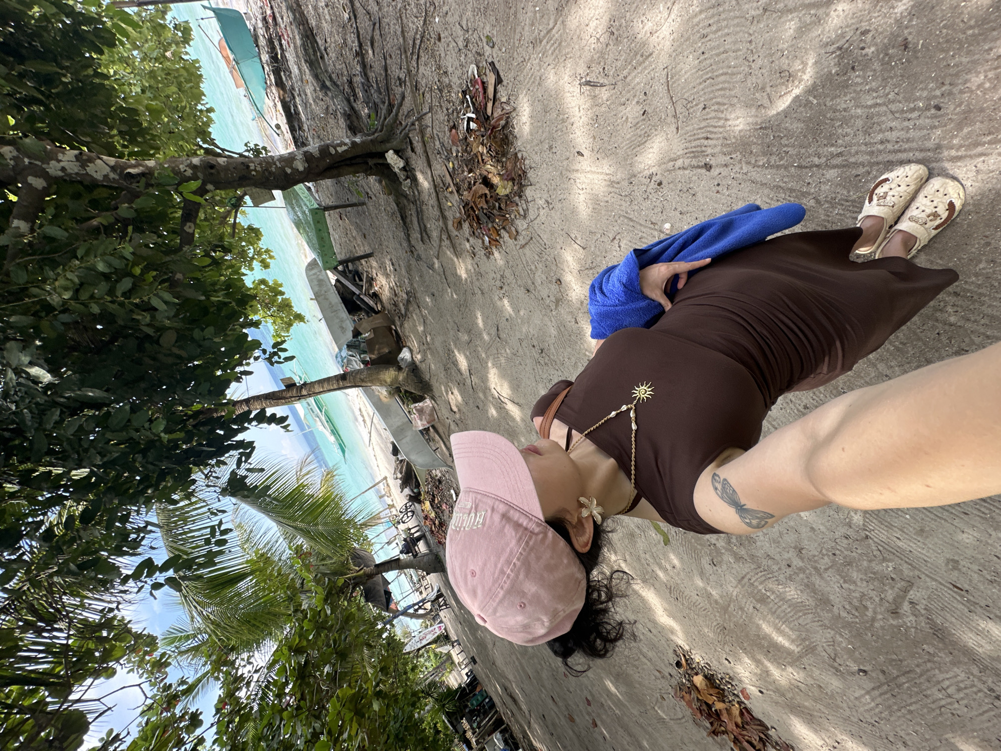 A woman wearing a pink baseball cap and a brown dress stands on a sandy beach. She has a tattoo of a butterfly on her left arm and is wearing a necklace with a sun pendant. She is holding a blue towel over her right arm. In the background, the turquoise water of the ocean is visible, with several small boats pulled up onto the shore. Palm trees and other vegetation line the beach, with debris and fallen leaves scattered on the sand. The scene suggests a casual day at the beach in Panglao, Philippines.