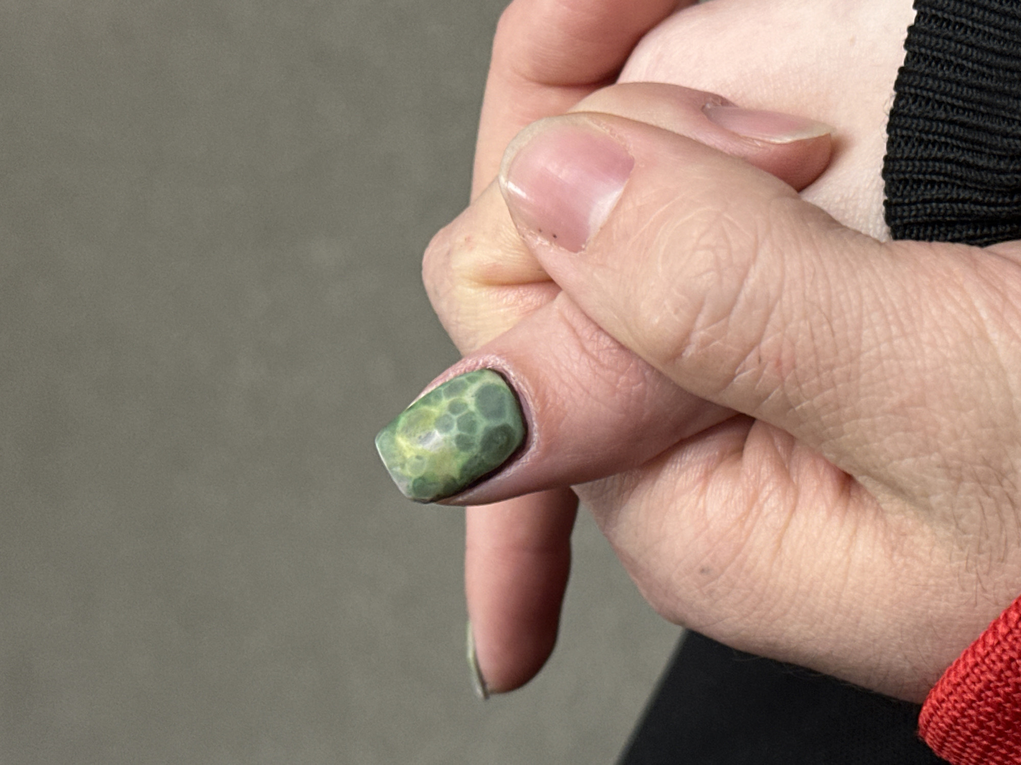 A close-up shot depicts a person's hand, with particular focus on the fingernails. The thumbnail is adorned with a green and yellow design, while the other visible fingernail is unadorned and natural. A portion of a black fabric and a red garment are also visible. There is no indication of this image being taken in Moscow, Russia.