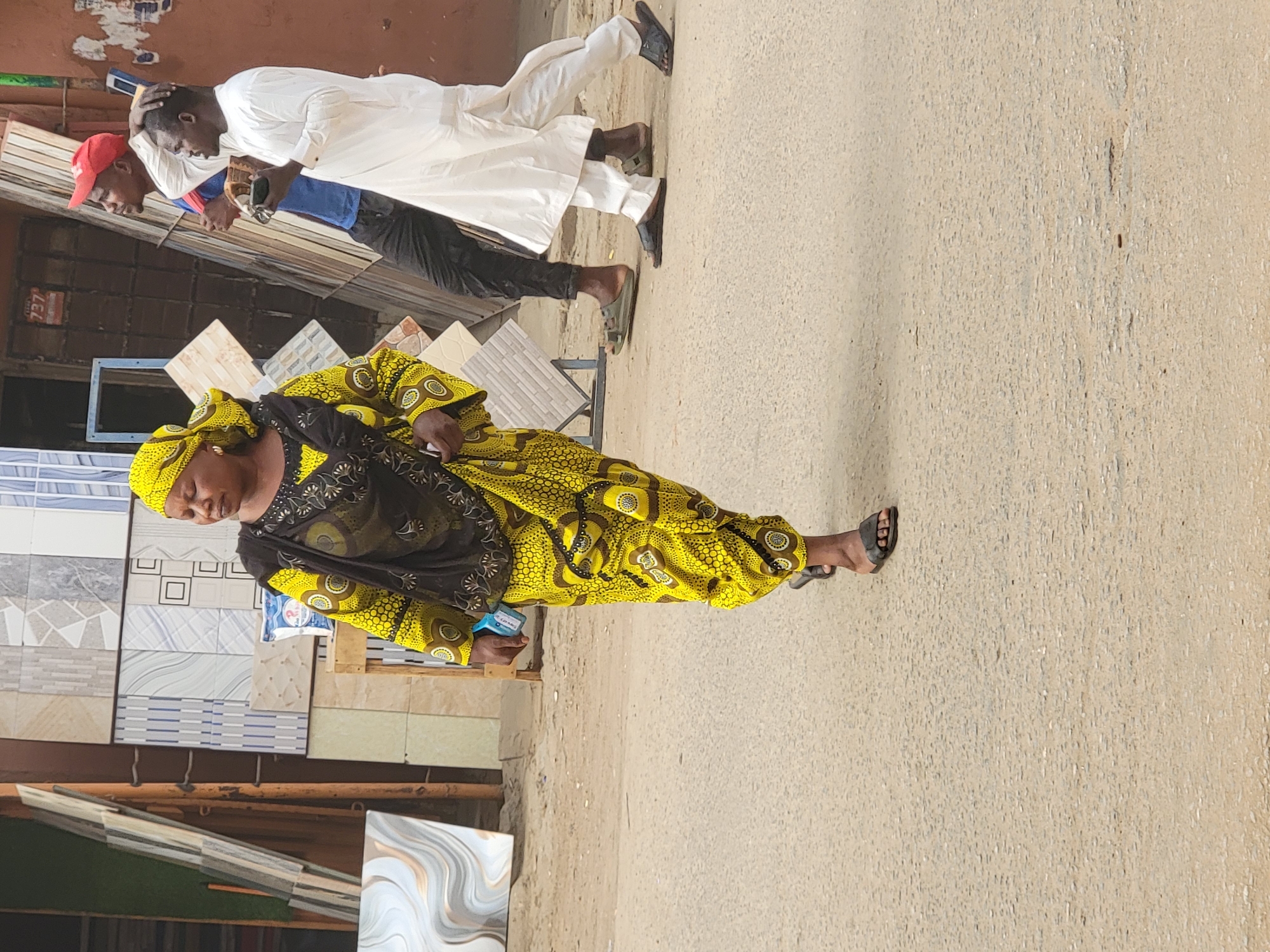 An outdoor scene in Maiburiji, Nigeria, depicts several people walking on a light-colored, dusty street or sidewalk, alongside a building materials shop displaying various wall and floor tiles. The image is brightly lit, indicating daytime.
In the foreground, a woman walks towards the right side of the frame, dressed in a vibrant yellow and black patterned long-sleeved garment and a matching yellow headwrap. She wears dark sandals and holds a small blue object in her left hand. Her gaze is directed slightly downwards.
Behind her, two men are also walking in the same general direction. The man closer to the viewer is wearing a traditional white long tunic (agbada-like) over dark pants and dark sandals or slippers. He holds a smartphone in his hands. Further behind him, another man wears a red cap, a blue shirt, and dark pants, also appearing to be engaged with a smartphone.
The background is dominated by displays of various ceramic and porcelain tiles, stacked and arranged along what appears to be a shopfront. The tiles feature a range of patterns, colors, and textures, suggesting a tile or building materials store. A small white label with "737" written on it is faintly visible on a dark surface behind the men. The overall setting is a busy street or market environment.