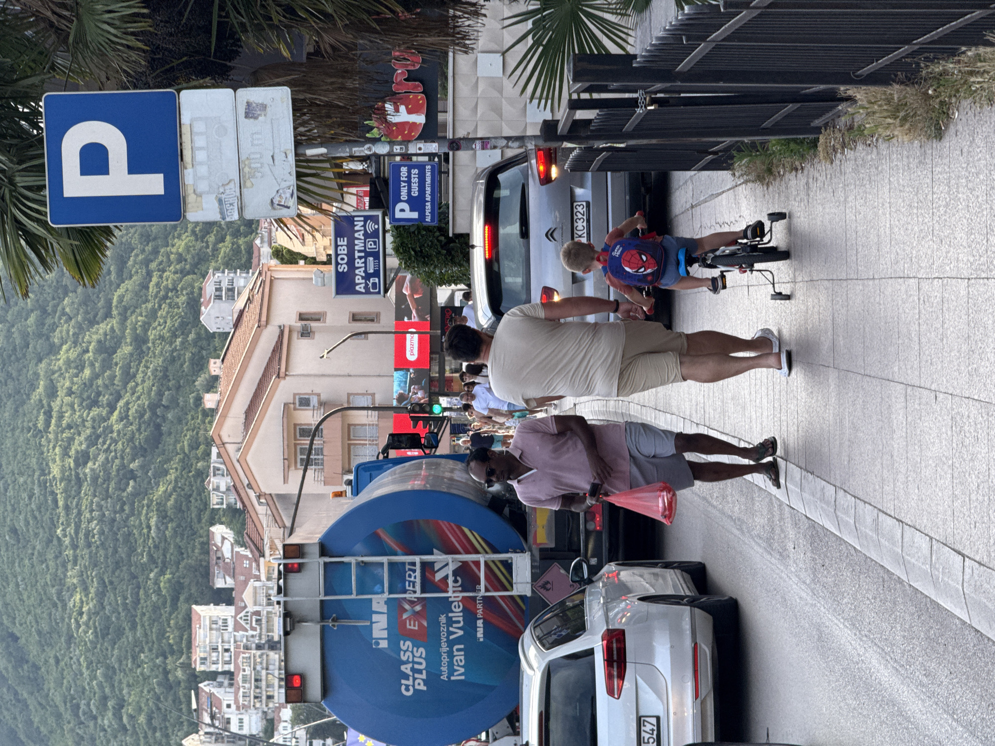 This image captures a bustling urban street scene in Budva, Montenegro, under daylight conditions.
In the foreground, a sidewalk made of grey paving slabs runs parallel to an asphalt road. On the sidewalk, a man in a light pink polo shirt, grey shorts, dark sunglasses, and dark flip-flops walks from right to left, carrying a translucent red plastic bag containing red items in his left hand and a dark smartphone in his right. Adjacent to him, another man in a light beige t-shirt, beige shorts, and white sandals walks from left to right, accompanying a young child. The child, wearing a red and blue Spider-Man backpack, blue shorts, and sneakers, rides a small red and black bicycle equipped with training wheels. A black metal fence separates the sidewalk from a strip of weeds and greenery on the far right.
On the road, traffic is present. A prominent blue tanker truck with "INA CLASS PLUS EXPERT Autoprijevoznik Ivan Vuletic INA PARTNER" in white and red lettering is visible, facing left. A hazard symbol (flammable liquid) is affixed to its rear. A white Volkswagen Passat Variant (B8) is positioned in front of the tanker, partially displaying the license plate "547". Further down the street, a grey Citroën C4 Picasso (second generation) is stopped, facing right, with illuminated rear lights and a license plate reading "KC 323".
Various signs are affixed to a pole on the right side of the sidewalk: a blue square sign with a white 'P' for parking, a faded sign depicting a bus and "500 m", and a smaller blue sign stating "P ONLY FOR GUESTS ALPESA APARTMENTS". Higher up, another blue sign reads "SOBE APARTMANI" (Rooms Apartments) with icons for TV, parking, and Wi-Fi. Palm fronds partially obscure some signs. In the background, multi-story buildings with light-colored facades and red/orange tiled roofs line the street, with additional pedestrians visible. A large, densely forested mountain forms the distant backdrop.