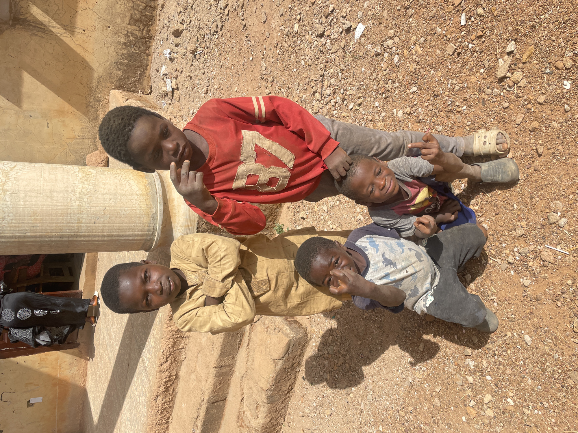 Four Black children are lying on the ground near a building. The environment appears to be Ladura Foron, Nigeria, as the area is characterized by a reddish-brown, rocky landscape.
One child is lying next to a white pillar and wearing a tan-colored shirt with long sleeves. Another child is wearing a red sweater with the number "87" on it, along with gray pants and mismatched shoes. A third child is wearing a gray shirt with a dinosaur pattern. The fourth child is wearing a gray long sleeved shirt with a pink and yellow design. Some are pointing at the camera.