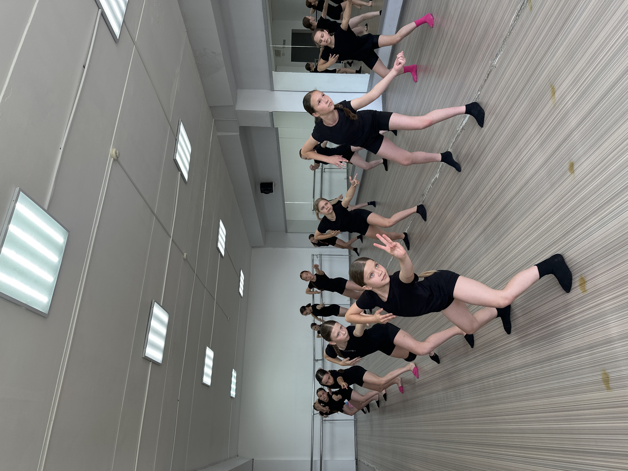 This image captures a dance class in session at a studio. A group of young girls, dressed in black leotards and shorts, are performing a choreographed routine. They are arranged in rows and are executing a variety of poses and movements with their arms extended and bodies angled. The studio appears to be a professional space with a mirrored wall along one side, ballet barres mounted on the wall, and a light-colored wooden floor. Overhead, rectangular fluorescent lights illuminate the room. The overall impression is one of a disciplined and energetic dance practice. The specific location of Votkinsk, Russia, is not discernible from the visual elements within the image.