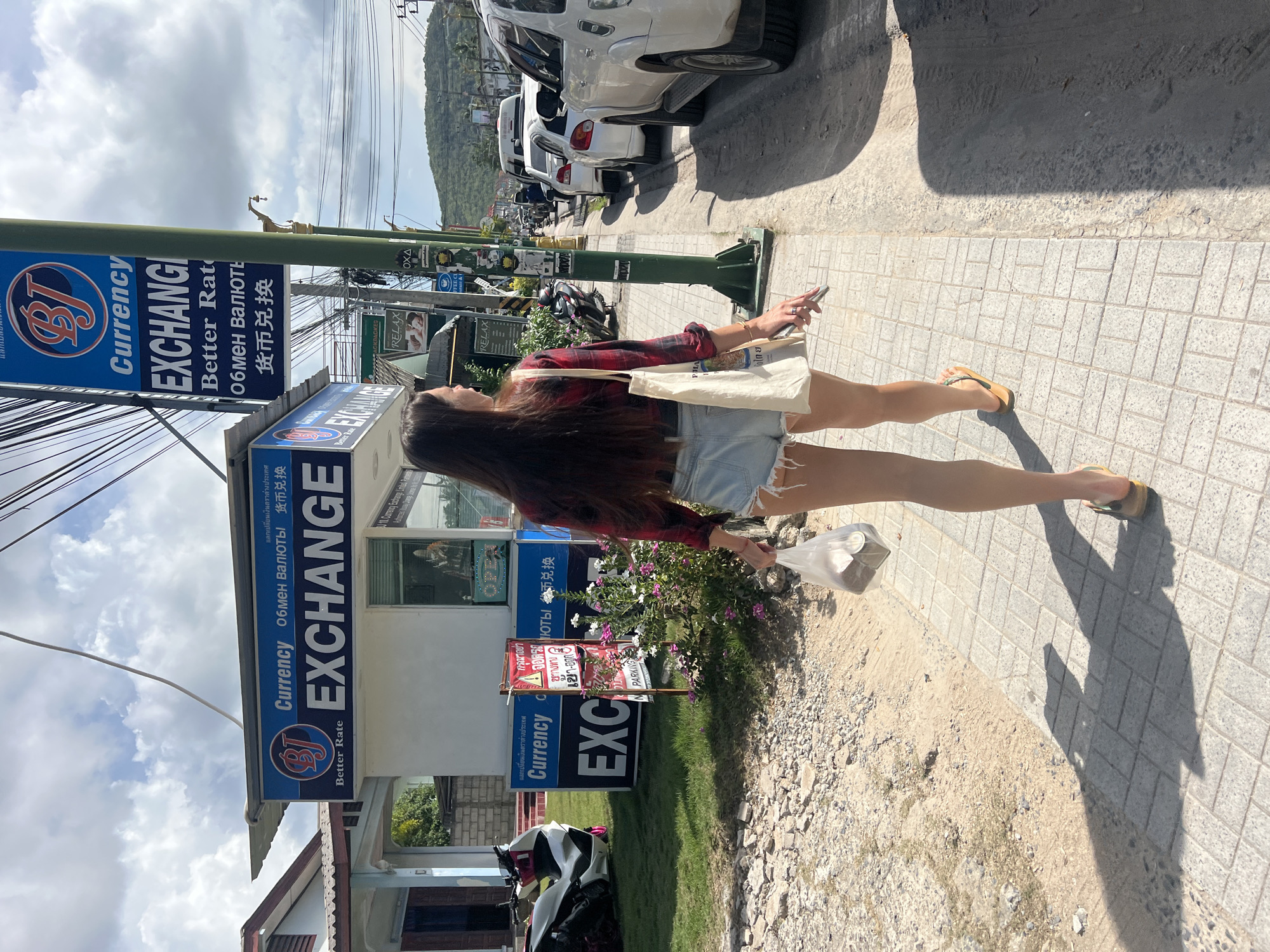 The image captures a moment on a street in Baan Bang Rak, Thailand. The primary subject is a woman with long, dark hair, walking on a paved sidewalk. She is wearing a red and black plaid shirt, denim shorts, and sandals. She holds a phone in one hand and a white tote bag in the other.
Beside her is a currency exchange kiosk with large, blue signage displaying "EXCHANGE" in English, Thai, Russian, and Chinese. Smaller signs with parking instructions are also visible.
Parked cars line the street. The sky above is partly cloudy. The general environment appears to be a commercial area, with overhead wires adding to the typical street scene.