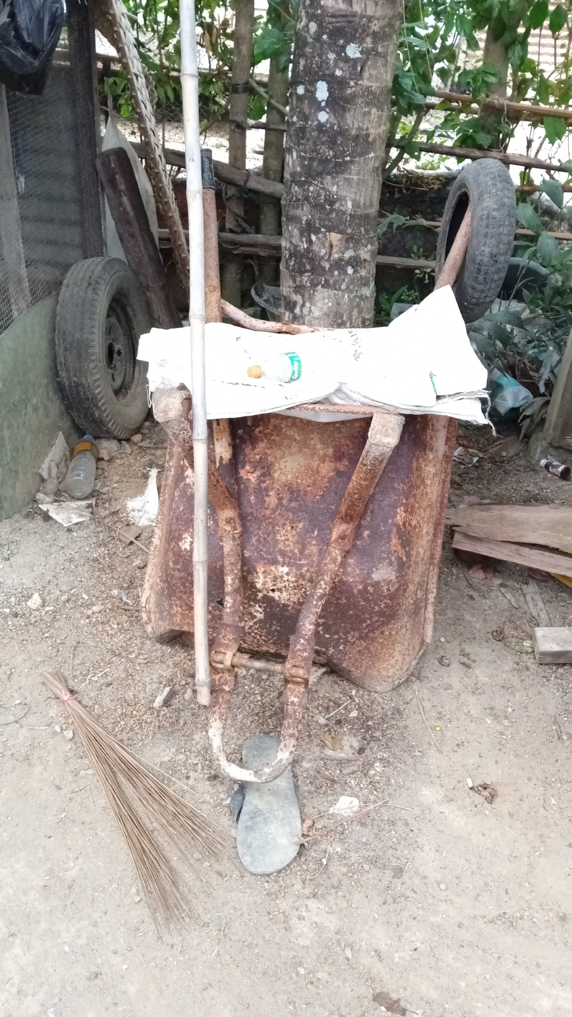 An exterior scene in Cotgadong, Indonesia, depicts a severely rusted wheelbarrow, likely an old cement mixer, filled with a white sack. A white plastic bottle with a green label and a brown cap rests on top of the sack. A grey flip-flop is placed on the ground in front of the wheelbarrow. To the left of the wheelbarrow, a broom made of dried grass is tied with red string and lies on the dirt ground. A large tree trunk with a rough bark texture stands behind the wheelbarrow, with a wooden fence and various other objects scattered around the area. The ground is a mixture of dirt, gravel, and debris. The overall impression is one of disuse and neglect.