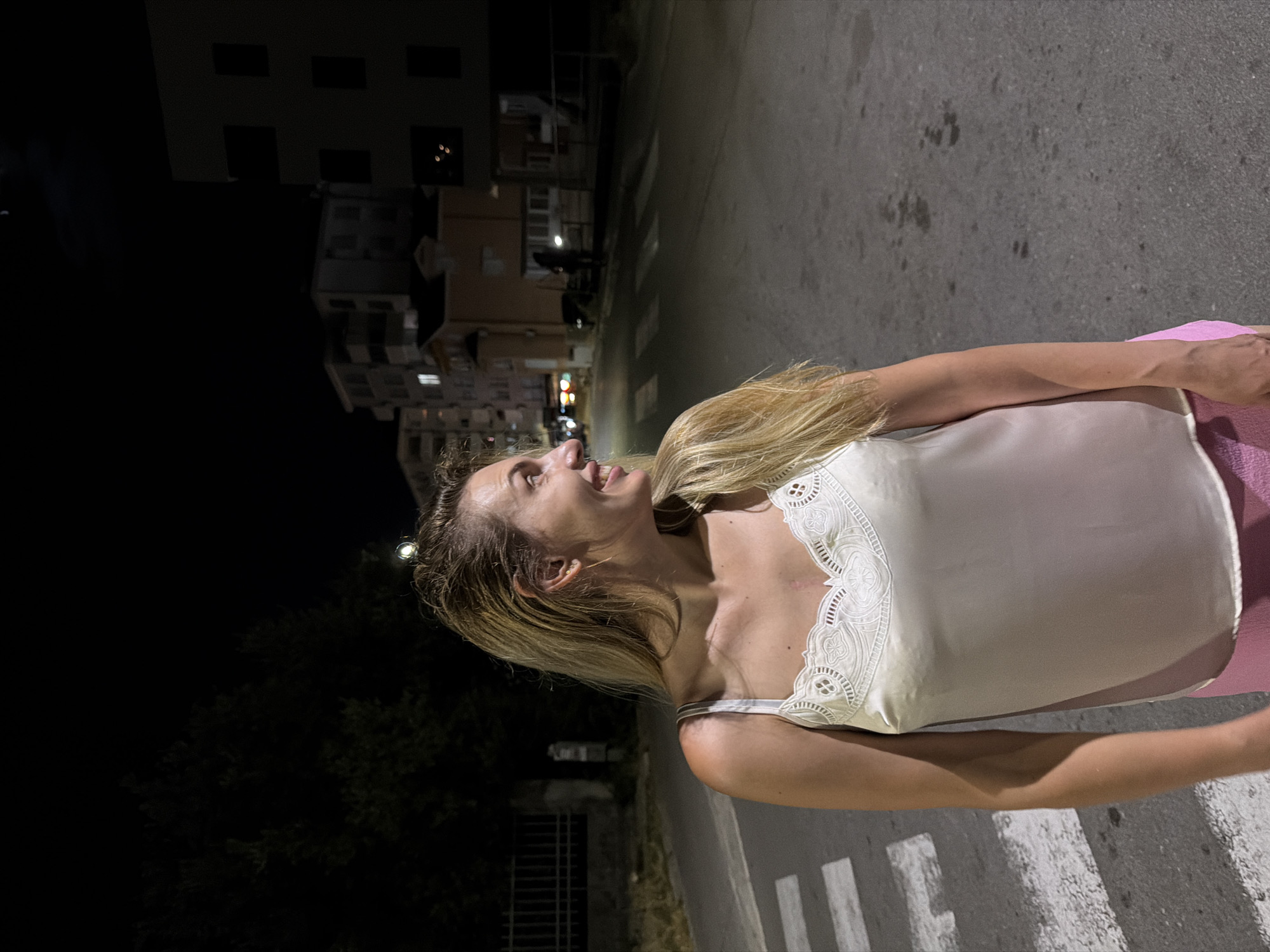 A blonde woman stands on a street at night in Budva, Montenegro. She is wearing a white tank top with lace trim and pink shorts. The woman is looking to her right, with a slight smile on her face. In the background, there are buildings, trees, and a street with a crosswalk. The moon is visible in the night sky.