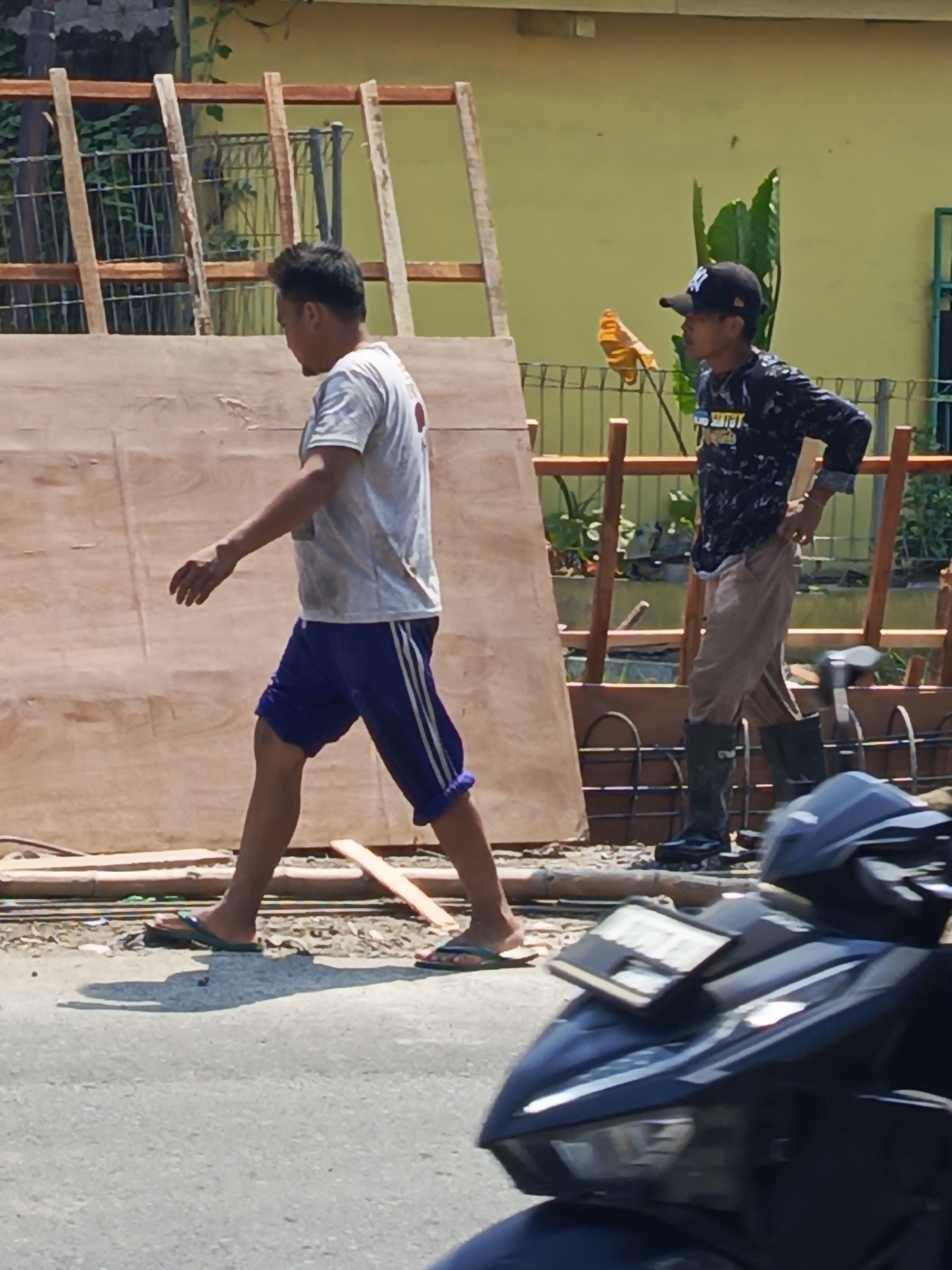 This image depicts a street scene in Kwangsan, Indonesia, on a sunny day. In the foreground, a dark blue motorcycle is partially visible, its front end angled towards the viewer. Two men are walking on a gravel path next to a large wooden panel supported by wooden scaffolding. The man on the left, wearing a light-colored t-shirt and blue shorts with white stripes, is walking towards the left of the frame, his body turned to the side. He is wearing green flip-flops. The man on the right, wearing a black patterned long-sleeved shirt, light brown pants, and black boots, is walking towards the right of the frame, his head turned slightly away from the viewer. He has a black baseball cap on. Behind the men and the wooden panel, a pale yellow wall is visible, with some green foliage and a metal fence in the background. The scene appears to be a construction or renovation area.