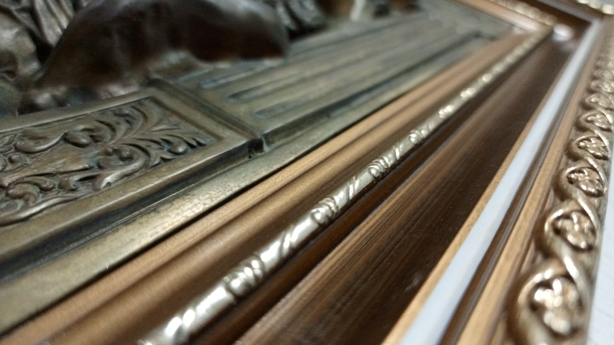 The image shows a close-up perspective of a framed artwork. The artwork itself features a bronze or brass relief with detailed floral and figurative designs. The frame is multi-layered and ornate, constructed of various wood and metal components. The innermost layer is a plain gold color, followed by a band with twisted rope detailing, then a dark wood section with a lighter wood trim, a white border, and finally an outermost layer with a repeating pattern of raised floral or leaf-like elements. The image does not provide any information about the location in the city of Los Robles, Venezuela.