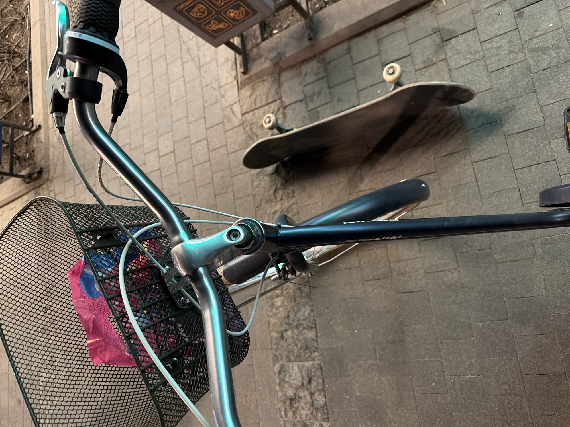 The image shows a bicycle, with a black wire basket attached to the handlebars, standing on a paved surface in a city. In the basket, there are partially visible items wrapped in pink and blue plastic. To the right of the bicycle, a skateboard lies on the ground. The surroundings consist of paving stones, possibly granite or a similar material. There is a metal structure, perhaps a signpost, situated in the background. The exact location in Moscow, Russia, cannot be determined from the given information.
