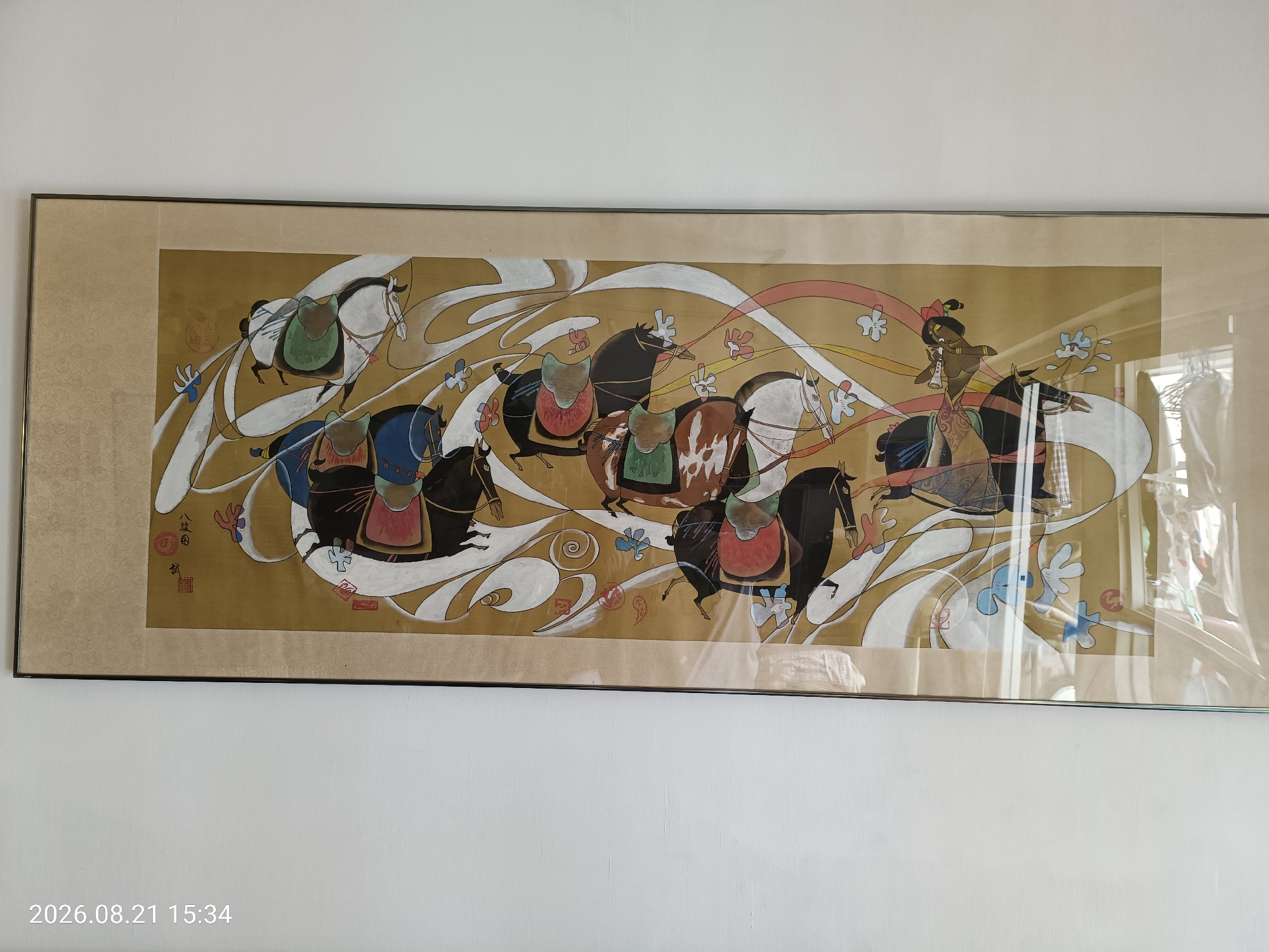 An unframed painting is displayed on a white wall. The painting is horizontal and depicts a procession of horses and riders. The horses are rendered in a stylized, almost cartoonish manner, with flowing lines and bold colors. Some of the horses are white, while others are black, brown, or a combination of colors. The riders are also stylized, with some wearing traditional Chinese clothing. The background of the painting is a muted gold color, with swirling white lines and small, colorful floral motifs. There are also some red and pink ribbon-like elements that appear to be in motion. In the upper left corner of the painting, there are two red seals. In the lower left corner of the image, a timestamp "2026.08.21 15:34" is visible. The image is taken from a slightly angled perspective, with some glare from what appears to be protective glass over the painting. The painting itself is the primary focus of the image, with the wall serving as a neutral backdrop. There are no discernible elements in the image that specifically indicate the location is Hong Kong, China, beyond the context provided in the prompt.