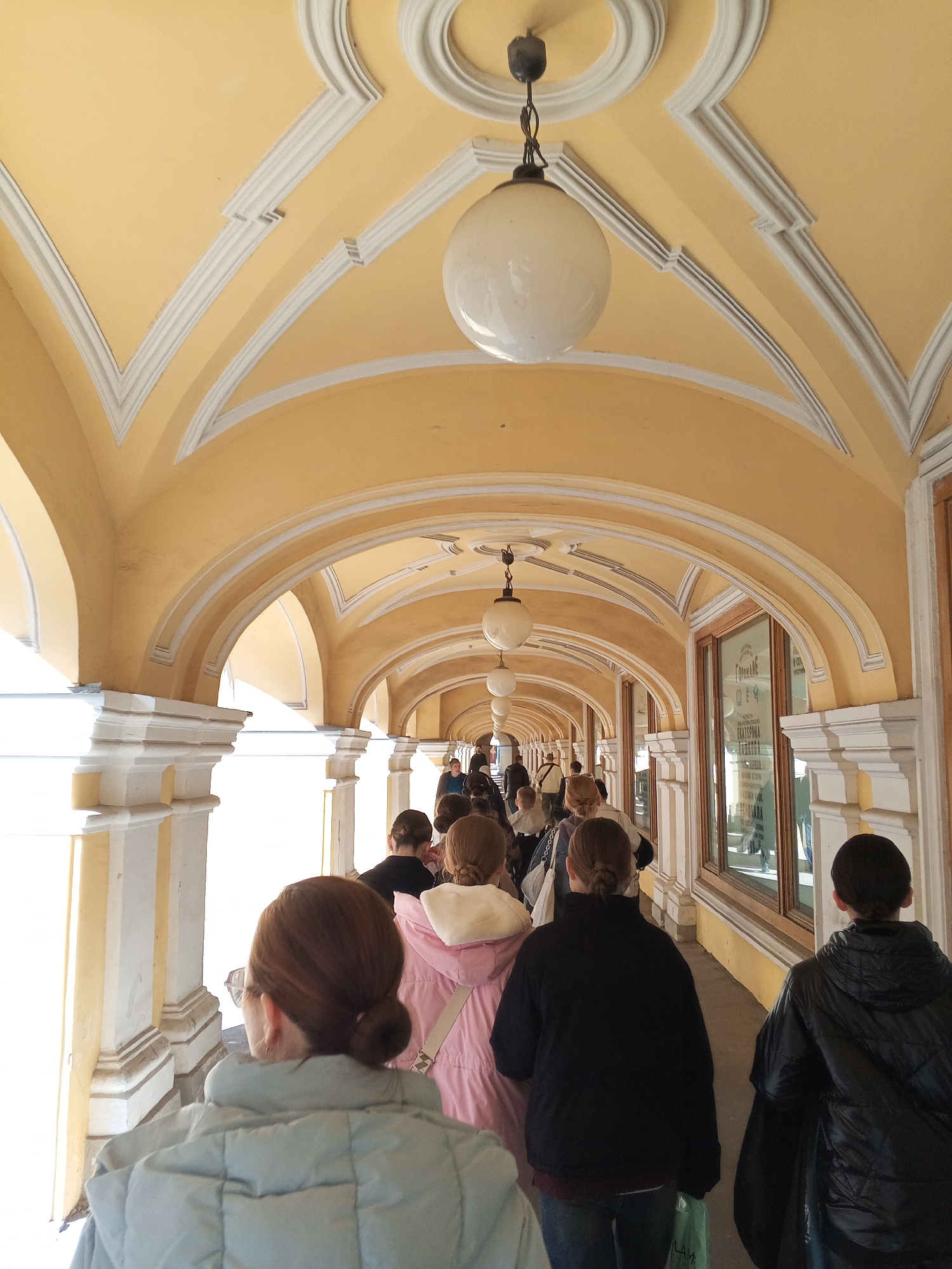 The photograph depicts a long, arched colonnade with people walking through it. The colonnade features a light yellow ceiling with white architectural details, including rounded arches and a circular design at the top of each arch. Spherical light fixtures hang from the ceiling. The colonnade runs along the side of a building with window displays visible on the right. People are walking away from the camera, their backs visible.
This location is in Saint Petersburg, Russia.