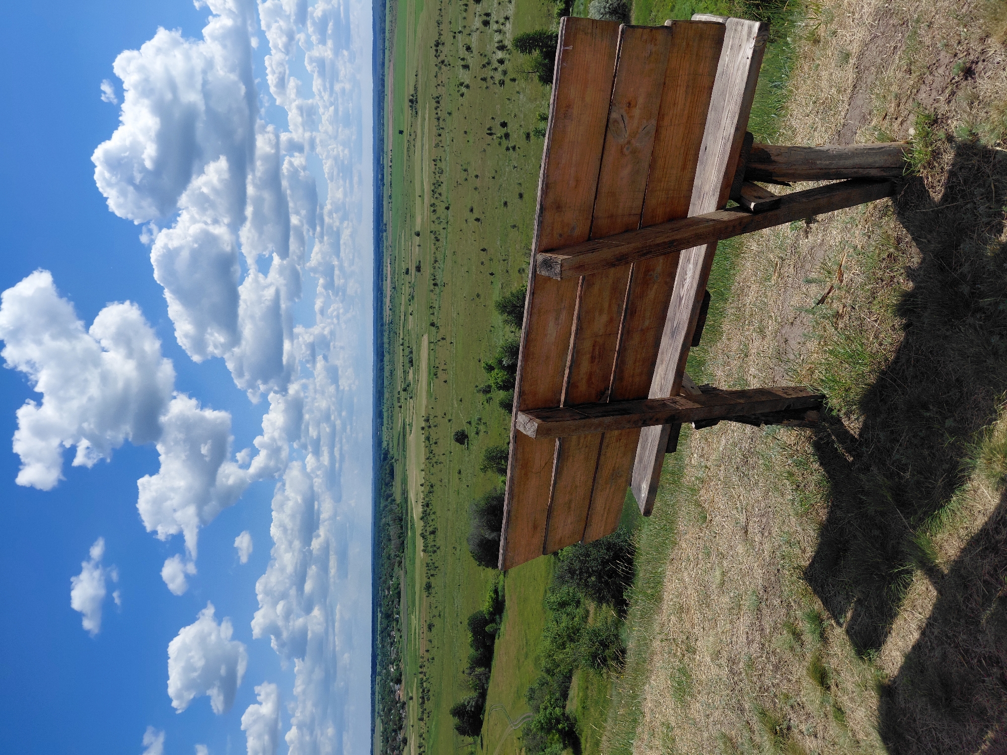 An outdoor shot captures a wooden bench positioned on a grassy hill overlooking a vast, green landscape under a partly cloudy sky. The bench is made of light-colored wood with visible grain and is supported by rustic wooden legs. Its backrest is a series of horizontal wooden planks. The foreground is dominated by dry grass and the shadow of the bench. The middle ground reveals a rolling expanse of green fields dotted with scattered trees and a distant village. The background features a hazy horizon line where the land meets a bright blue sky filled with fluffy white clouds. The scene appears serene and rural.