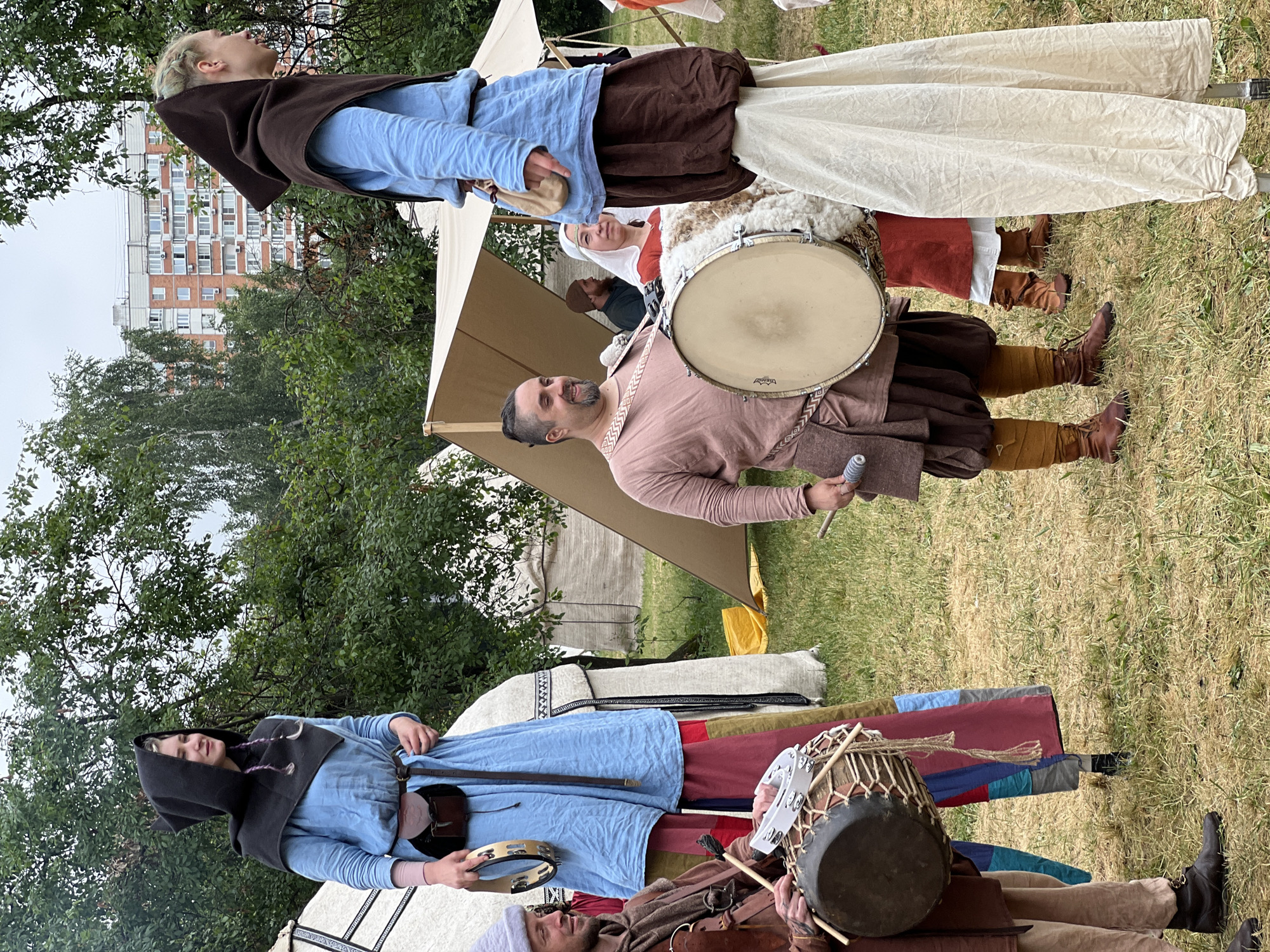The image shows an outdoor historical reenactment event. In the foreground, two individuals on stilts wear medieval-style costumes. The person on the left is dressed in a blue tunic and dark hood, holding a tambourine. The person on the right wears a blue tunic and a brown hood, with a brown skirt and a cream-colored underskirt. To the left of the stilted figures, a man in a brown tunic and cap is playing a drum while holding a tambourine. Behind him, another man with a beard in a green tunic is partially visible. A woman in a red tunic and white headscarf is seated between the stilted figures and the drummer. In the background, tents and trees are visible, with a modern apartment building in the far distance. The ground is covered in grass and dried vegetation. This scene is likely taking place in a park or open space within the city of Moscow, Russia, given the presence of a contemporary urban building in the background.