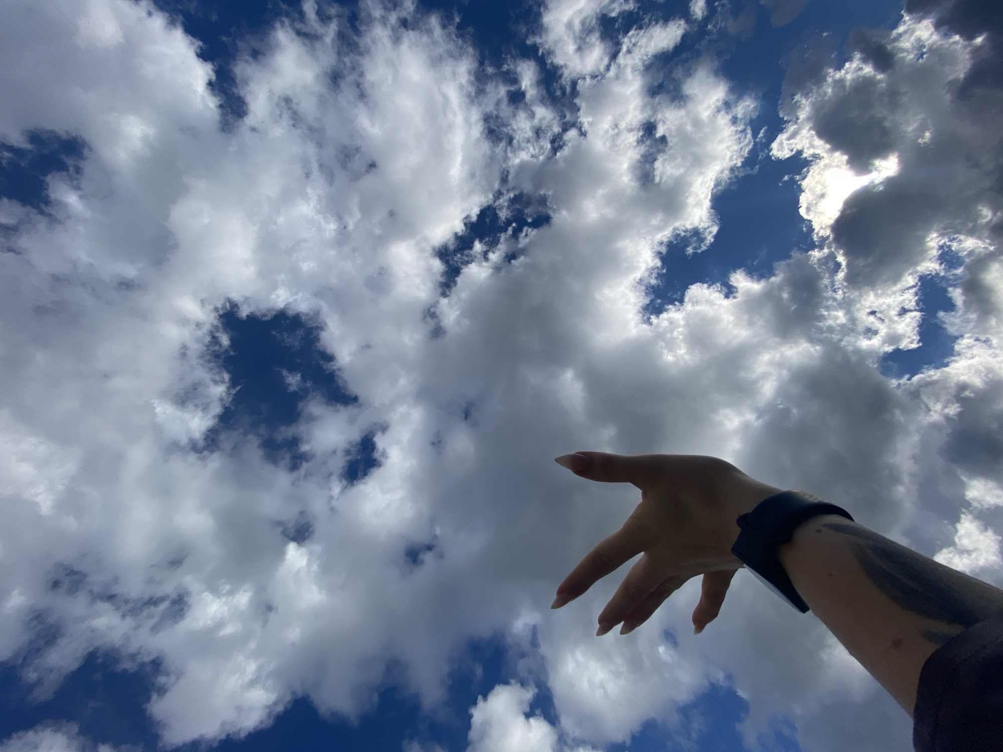 The image displays a human arm and hand, seen from below, extending upwards and slightly to the left against a backdrop of a vibrant blue sky filled with clouds. The hand is open with fingers spread, appearing to reach towards the sky. The nails are long and manicured. A dark-colored smartwatch or fitness tracker is worn on the wrist. A prominent, dark-toned tattoo with abstract, possibly organic or swirling designs, is visible on the forearm.
The sky exhibits a mix of bright white and light grey clouds, with patches of clear, deep blue visible between them. On the upper right side of the frame, the sun's brilliance is partially obscured by clouds, creating a strong glare and radiating light, indicating a bright, sunny day with significant cloud cover.
The perspective from beneath the arm emphasizes the vastness of the sky. The lighting suggests it is daytime, likely mid-day, given the intensity of the sunlight filtering through the clouds. The scene is set in Chaikovsky, Russia. There is no visible text in the image.