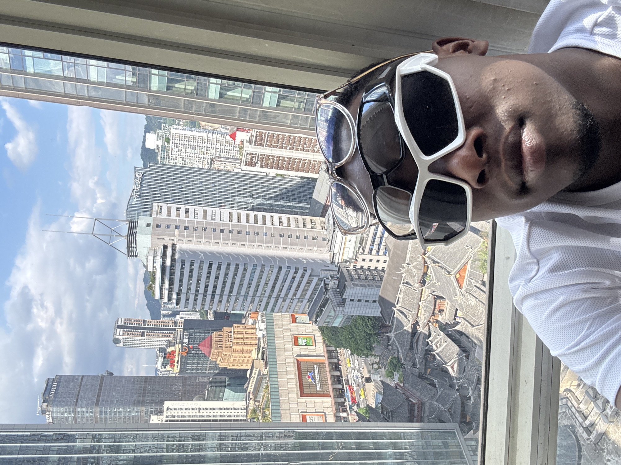 A selfie taken from a high-rise window in Yuzhong District, China, shows a person's face in the foreground, wearing two pairs of sunglasses. The person is wearing a white polo shirt. In the background, a sprawling cityscape unfolds, featuring numerous skyscrapers and traditional Chinese architecture. A large shopping mall with red banners is visible in the mid-ground, along with a dense cluster of gray-roofed buildings that suggest an older district. The sky is blue with scattered clouds. The overall impression is of a modern metropolis with preserved historical elements.