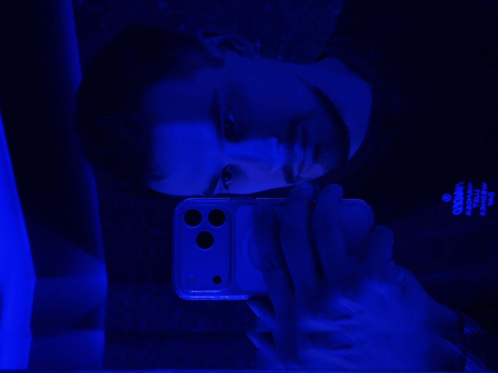 A dark and moody image, heavily saturated with blue light, captures a person taking a mirror selfie. The central subject is a young man, reflected in what appears to be a large mirror, with his head tilted towards the viewer. He has short hair and a subtle beard, and his eyes are directly engaging the camera.
He is holding a smartphone, specifically an iPhone with a triple-camera array and a clear case featuring a MagSafe ring, in his right hand. His hand is visible, partially obscuring the phone, as he positions it for the selfie. The blue illumination casts deep shadows and highlights across his face and hand, emphasizing the contours and adding to the dramatic atmosphere.
The setting appears to be indoors, likely at night or in a low-light environment designed with blue lighting, possibly a club, bar, or a themed room in Krasnoyarsk, Russia. The overall dark and blue aesthetic creates a mysterious and cool ambiance.
Visible on the man's shirt, reflected and rotated in the mirror, is text. When corrected for mirroring and rotation, the text reads "MAXO WASHAW MEDWED YAG". The word "MEDWED" (bear) is notably decipherable. No specific emotions are clearly discernible beyond a neutral, direct gaze. The image is presented at a 90-degree clockwise rotation, adding an unusual perspective to the reflection.