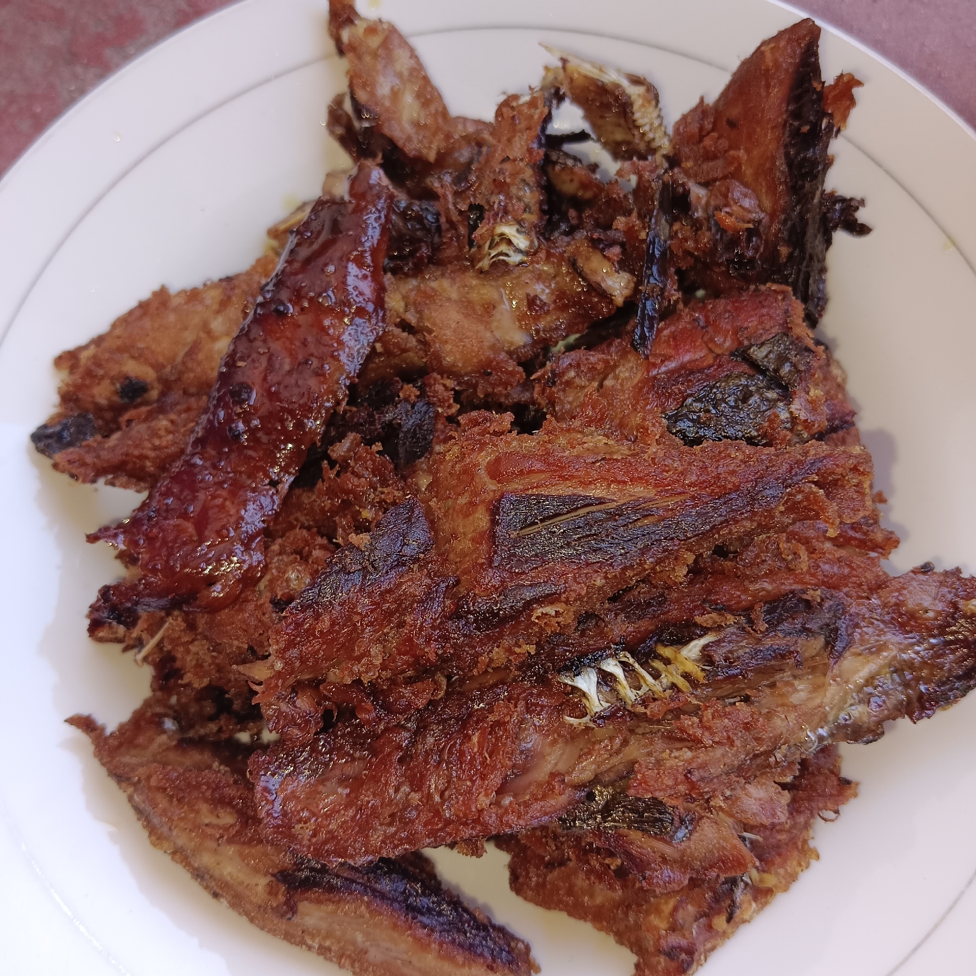 A plate of fried fish is presented, likely in a home setting in Paupanda, Indonesia. The fish appears to be small and whole, with a crispy, golden-brown exterior. Some pieces are coated in a glistening sauce, possibly soy sauce or a sweet and savory glaze. The fish is piled high on a white plate, which is resting on a textured reddish surface, possibly a tablecloth or placemat. The lighting suggests an indoor environment, with soft, diffused light casting subtle shadows. The overall impression is of a home-cooked meal, ready to be enjoyed.