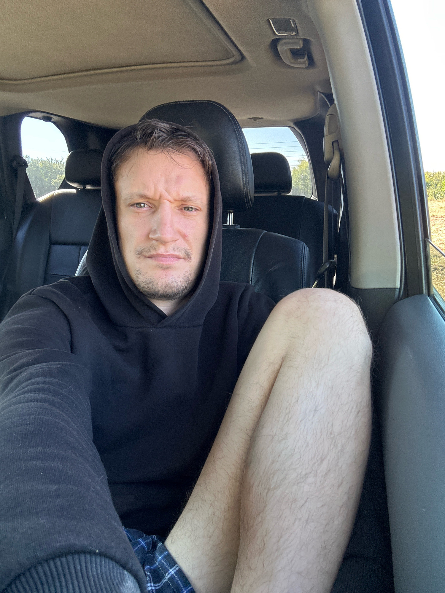 A medium shot shows a man with light brown hair and stubble sitting in the driver's seat of a vehicle. He is wearing a black hooded sweatshirt and plaid shorts. His left leg is raised and bent, with his knee pointing towards the camera. The interior of the vehicle is visible, including black leather seats and a sunroof. Outside the vehicle, there is a bright sky and greenery. The image appears to have been taken during the daytime. The location is not discernible from the image.