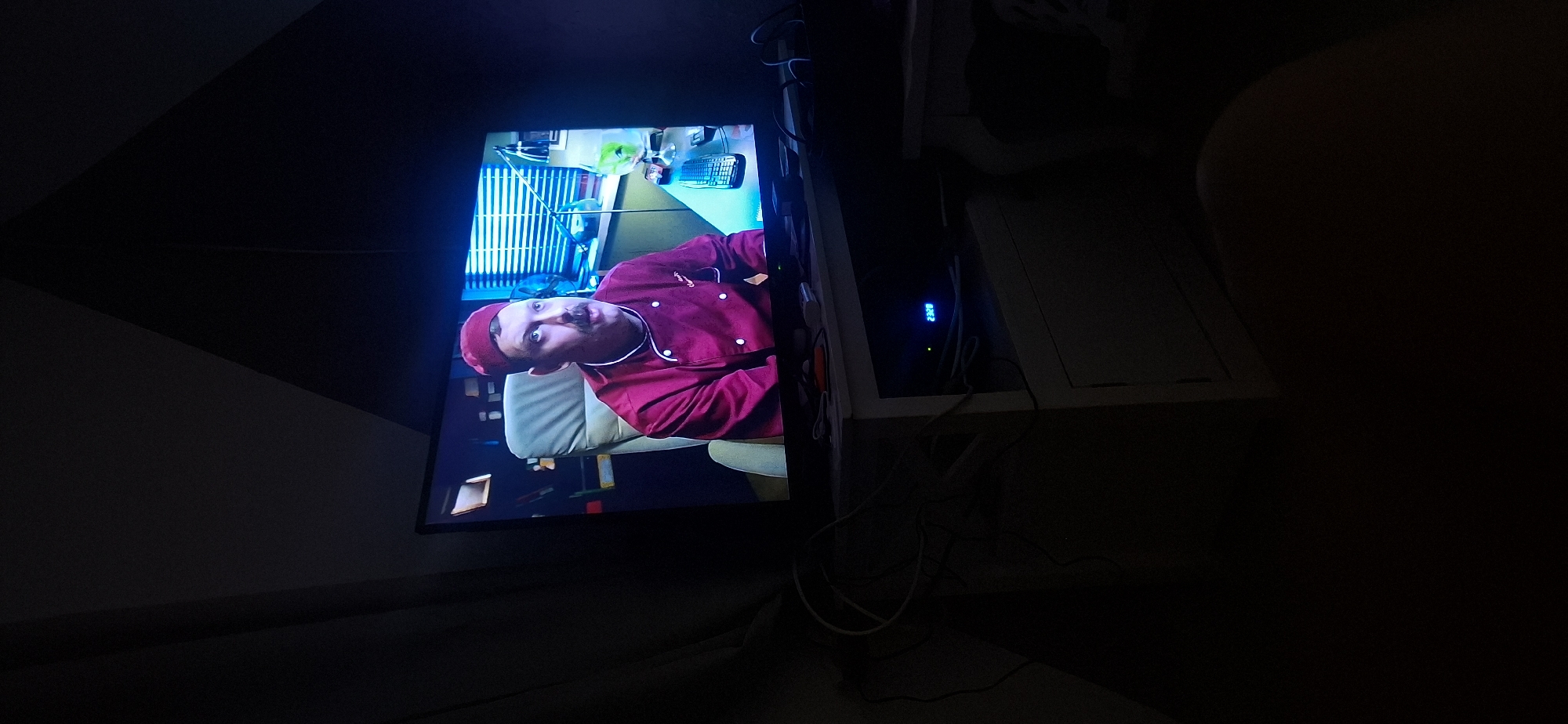 A man wearing a red chef's uniform and a matching hat, with a prominent mustache, is seated in a swivel chair in front of a television screen. The television displays the man's image, suggesting it's a recording or broadcast. He appears to be looking towards the right side of the frame. The background on the screen shows a desk with blinds, a fan, and a green drink in a stemmed glass. In the foreground, the television is placed on a white cabinet. The cabinet has a digital display showing "23:28." Cables are visible on and around the cabinet, and the room is dimly lit, with dark curtains on the left side of the image. The image appears to be taken in a residential setting in Yerocham, Israel.