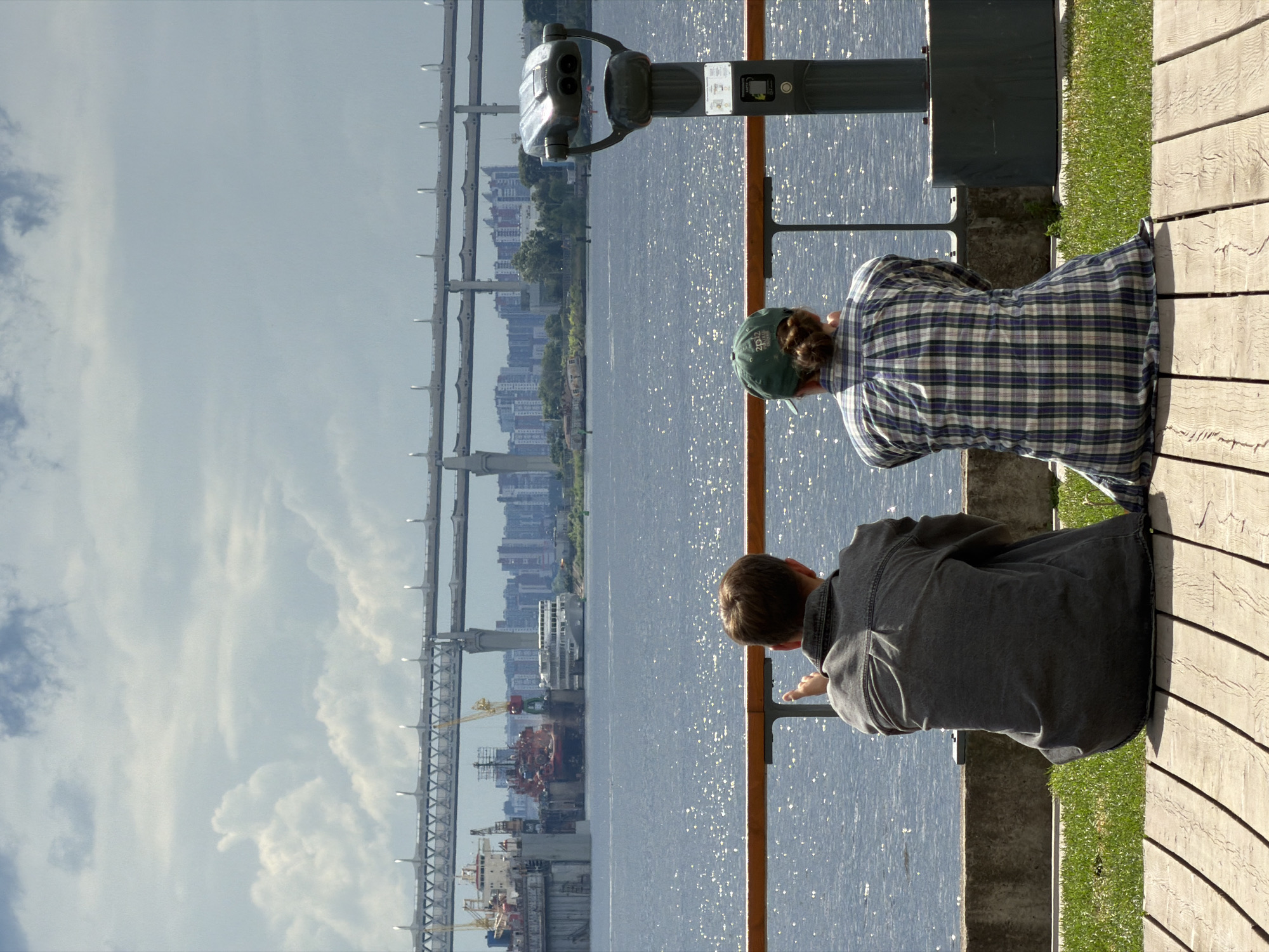 Two individuals are seated on a wooden deck, facing a body of water. The person on the left, wearing a grey denim jacket, is pointing towards the water. The person on the right, wearing a green baseball cap and a plaid shirt, is looking out at the same view. In the background, a large bridge spans the water, with ships docked in a shipyard to the left. The city skyline is visible on the horizon. A large, coin-operated viewer is positioned on the right side of the deck. The scene is set on a cloudy day with sunlight glinting off the water. This appears to be a scenic viewpoint along the Neva River in Saint Petersburg, Russia, possibly near the Western High-Speed Diameter.