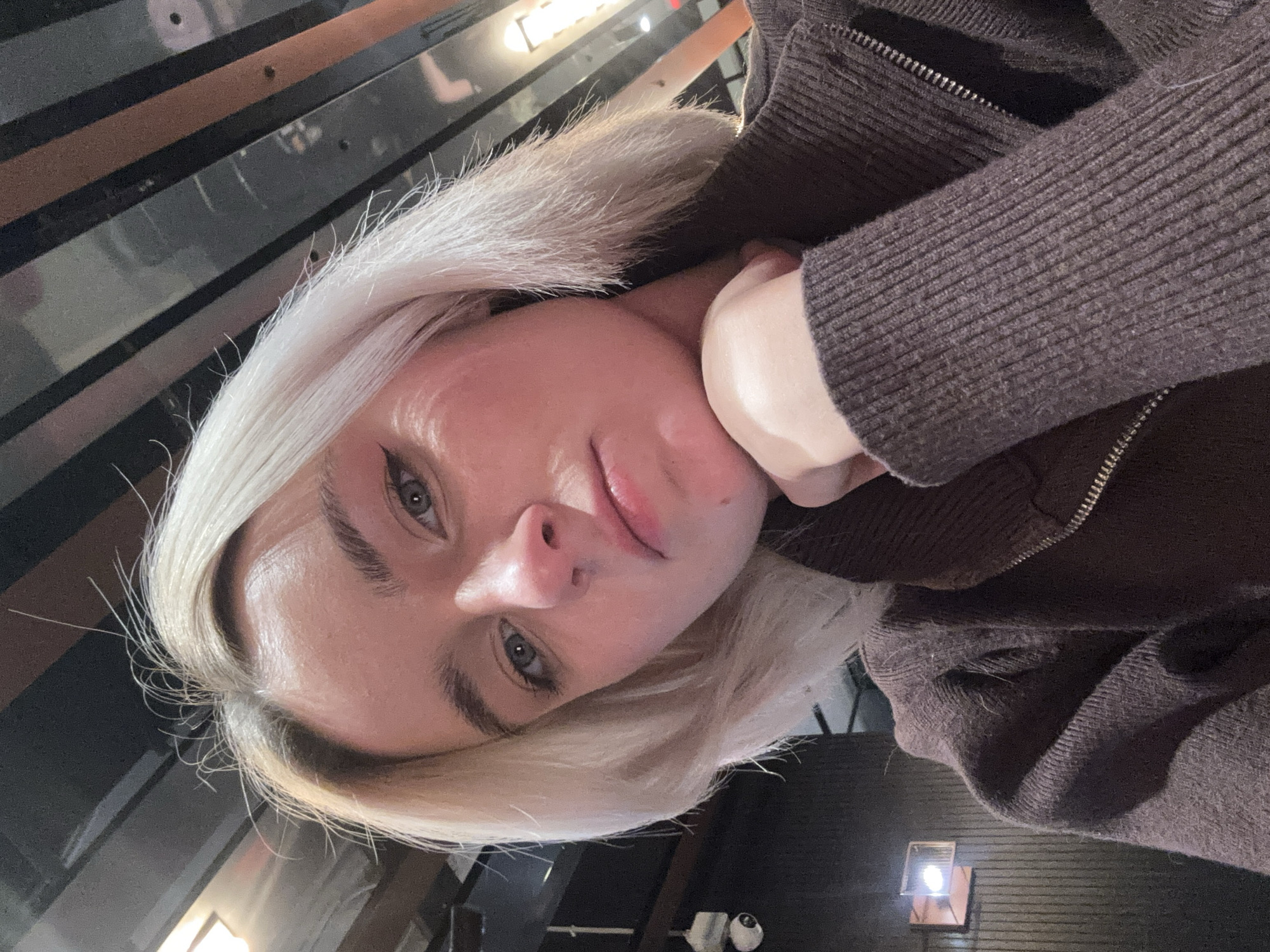 A selfie-style, angled close-up depicts a fair-skinned person with dyed platinum blonde hair, wearing a brown, zipped sweater or jacket. The person is resting their chin on their hand, looking directly at the camera. The background appears to be an interior space, possibly a restaurant or bar, with dark horizontal lines and a light fixture visible. The location is not discernible as Sinyavino, Russia, from the provided image.