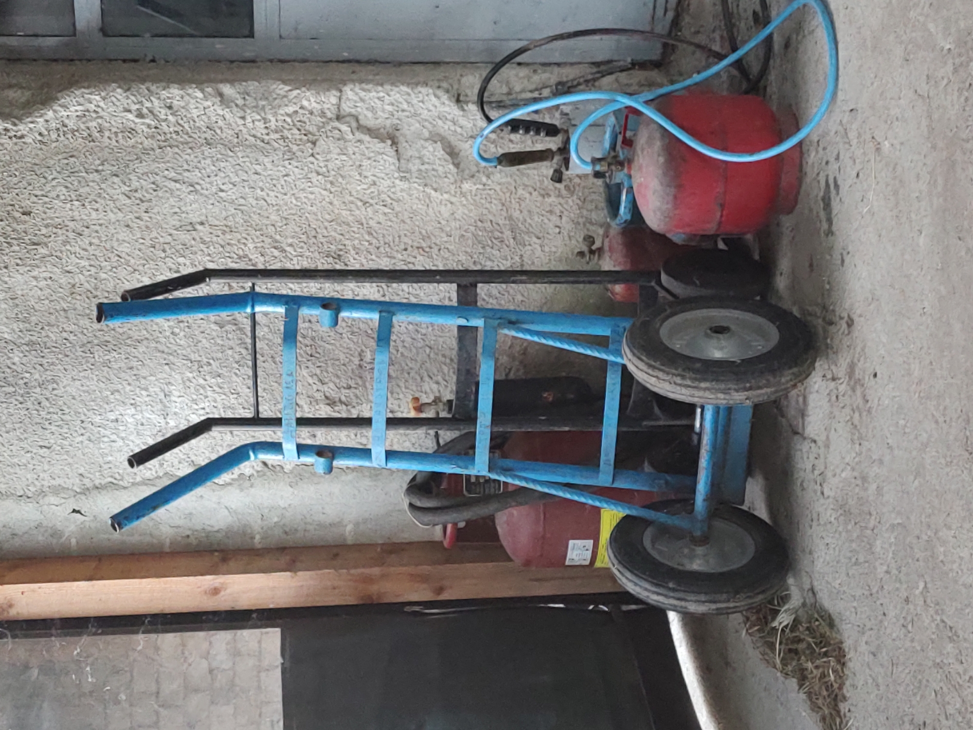 The image shows a blue metal cart with two wheels, possibly a dolly or hand truck, parked next to a red air compressor tank. The cart has black handles at the top. The air compressor tank is round and cylindrical, and a blue hose is connected to it. It appears to be an outdoor or workshop setting, with a rough textured wall in the background and a wooden shelf or beam below the cart. The lighting suggests it is daytime. There is some dry grass or straw on the ground near the wheels. The scene is static, with no activity taking place. There is no visible text in the image. The location is identified as Kochmar, Bulgaria.