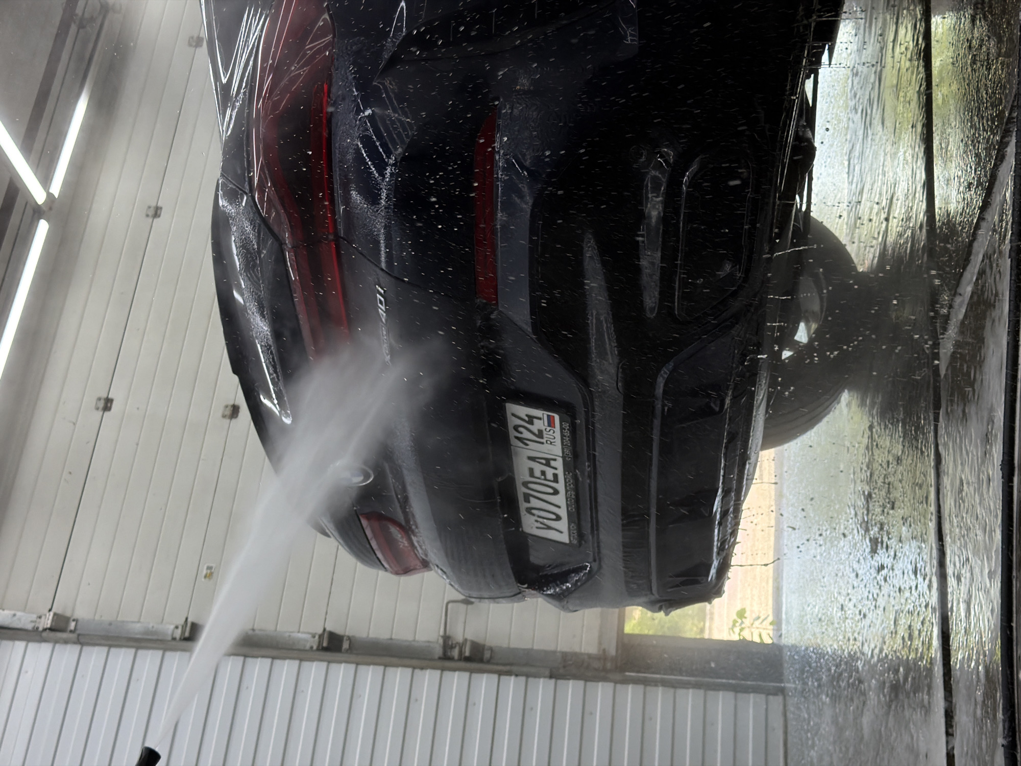 In Krasnoyarsk, Russia, a dark-colored car is being washed with a high-pressure water spray in an indoor car wash bay. The rear of the vehicle, including its taillights and license plate, is visible. The license plate reads "Y070EA 124 RUS". Water is spraying across the back of the car and onto the wet floor. The bay appears to have a corrugated metal wall and fluorescent lighting overhead. The overall scene depicts a typical car washing process.