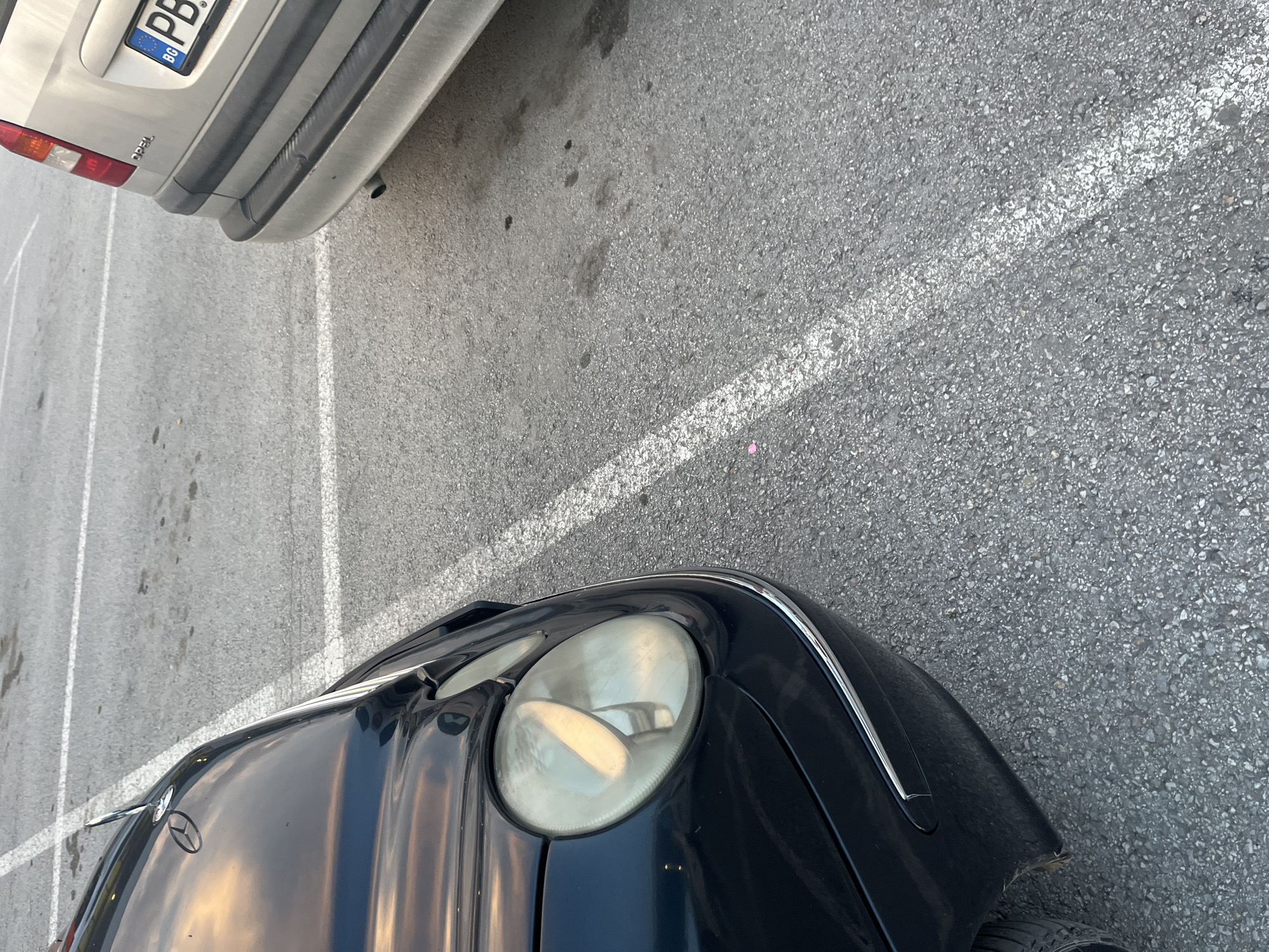 This media file is a top-down, close-up shot of two cars parked on an asphalt surface, likely a parking lot. The lighting suggests it is daytime, possibly late afternoon, with some shadows cast by the vehicles. The asphalt is grey and textured, with white painted parking lines visible.
The foreground features the hood and headlight of a dark-colored Mercedes-Benz. The Mercedes emblem is clearly visible on the hood, and the headlight is large and round, showing reflections of the sky. The car appears to be in good condition, though there's a slight scuff on the front bumper.
In the background, the rear of a lighter-colored car, possibly an Opel, is visible. A license plate with the letters "PB" and the country code "BG" (indicating Bulgaria) is clearly readable. The rear bumper and a taillight are also visible.
The scene appears to be a standard parking area, with no people or other significant activity. The focus is on the vehicles and the ground. The location context provided is Plovdiv, Bulgaria, which aligns with the "BG" on the license plate. The overall impression is one of urban automotive parking.