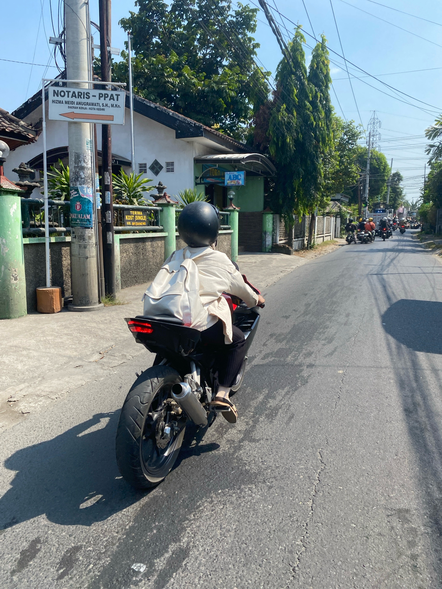 The image depicts a street scene in the city of Kediri, Indonesia. In the foreground, a person on a motorcycle is seen from behind, wearing a black helmet and a light-colored jacket, with a white backpack on their back. The motorcycle has a black body and a silver exhaust pipe. The road surface is asphalt with visible cracks and some debris. On the left side of the road, there is a pole with several signs, one of which reads "NOTARIS - PPAT HIZMA MEIDI ANUGRAWATI, S.H., M.Kn." with an arrow pointing to the right, indicating the direction to the notary office. Further down the street, a procession of other motorcycles with riders is visible, suggesting a group activity or a common mode of transportation in the area. Tall trees line the right side of the road, and power lines crisscross the sky. The buildings visible appear to be single-story structures with some green foliage in front. The overall environment suggests a sunny day in a residential or commercial area.