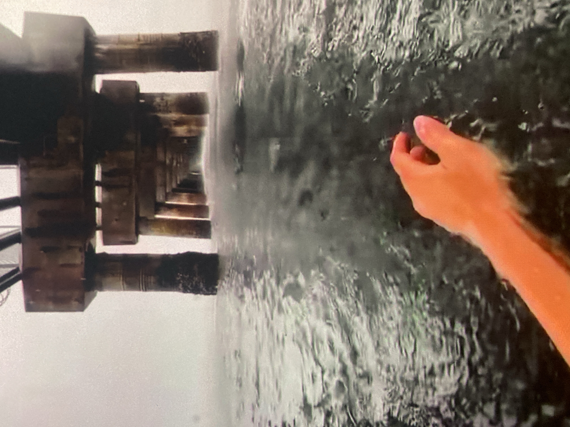 The image shows a person's hand reaching out into choppy water under a pier. The pier is made of concrete pillars and beams, and it extends into the distance. The water is dark and reflects the pier above. The sky is overcast and hazy. The scene appears to be outdoors, possibly at a beach or a harbor. The specific location in the city of Ustyvytsia, Ukraine, is not discernible from the image.