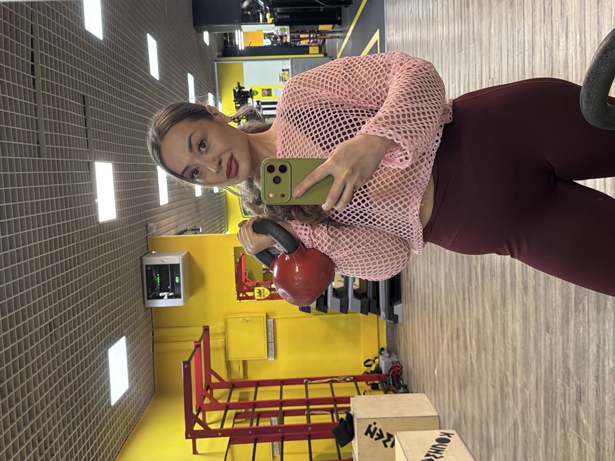 The image, rotated 90 degrees clockwise, captures a woman taking a selfie in what appears to be an indoor fitness facility or gym in Krasnoyarsk, Russia. The woman, with light brown hair pulled back, looks directly into the camera of her green-cased smartphone, which she holds up. She wears a light pink, long-sleeved mesh top over a dark top, and high-waisted maroon or dark red leggings. Her left hand holds the black handle of a red kettlebell, suggesting she is mid-workout or posing in a gym context.
The background reveals a dynamic gym environment. The floor is made of light-colored wooden planks. The walls feature distinct sections: one side has a light gray, textured grid pattern with bright rectangular lights embedded, while another section is painted a vibrant yellow. In the yellow area, there's a red metal rack or squat stand, and various pieces of dark gym equipment are visible further back. A small, light gray electronic device or monitor is mounted on the yellow wall. Near the bottom of the frame, several light brown wooden plyometric boxes are stacked, with some visible text, possibly "KOUKRA," on their sides. The lighting is bright and even, indicating an indoor setting.