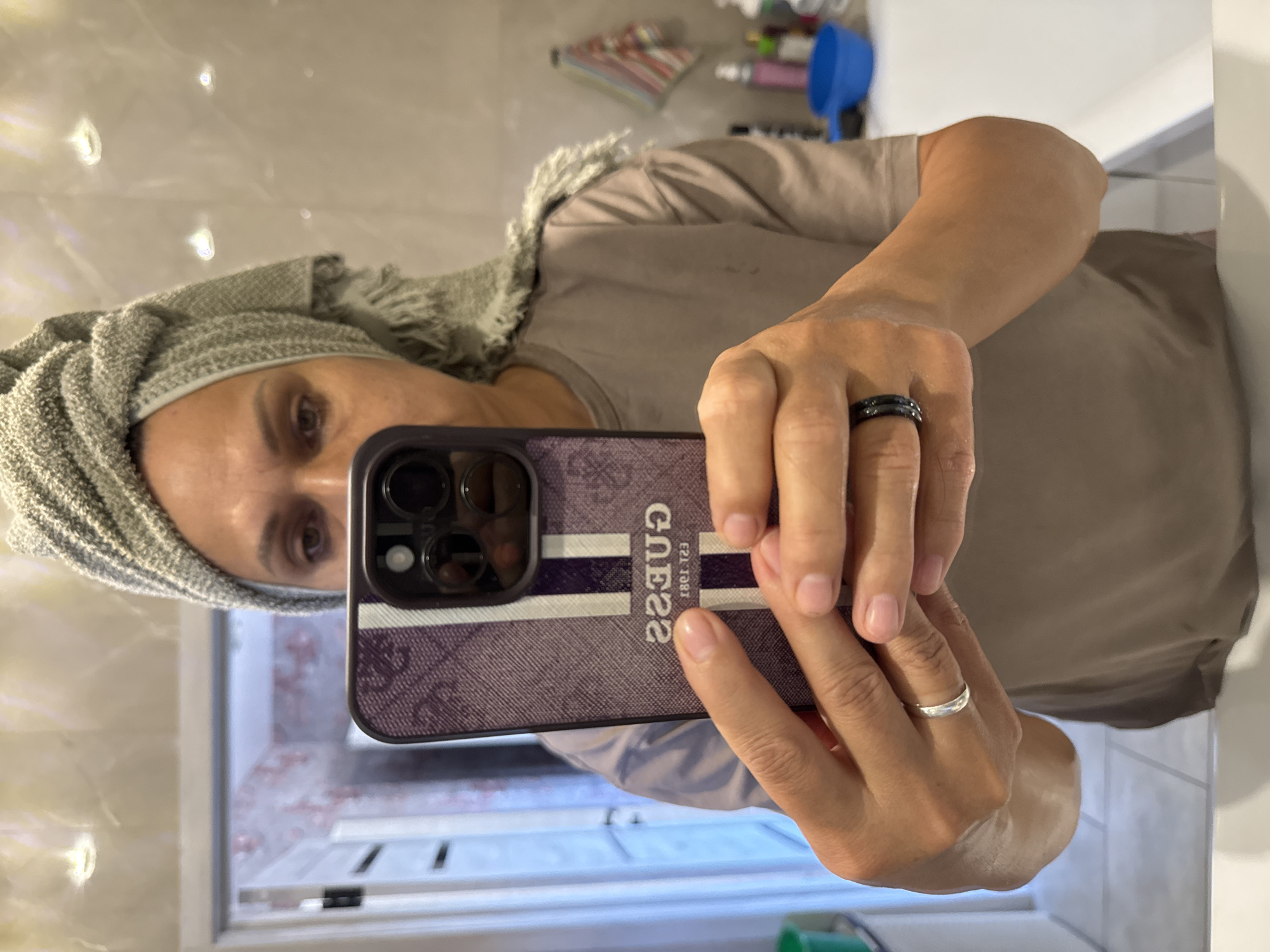The image shows a selfie taken by a woman in a bathroom. She is wearing a grey towel turban on her head and a beige t-shirt. She is holding a smartphone in her hands, which has a purple and white striped case with the word "GUESS" printed on it. The woman is wearing a silver ring on her left hand and a black ring on her right hand. The bathroom has tiled walls and a mirror. There are some toiletries visible in the background, including bottles and a blue bowl. The location is the city of Soloneshnoye, Russia.