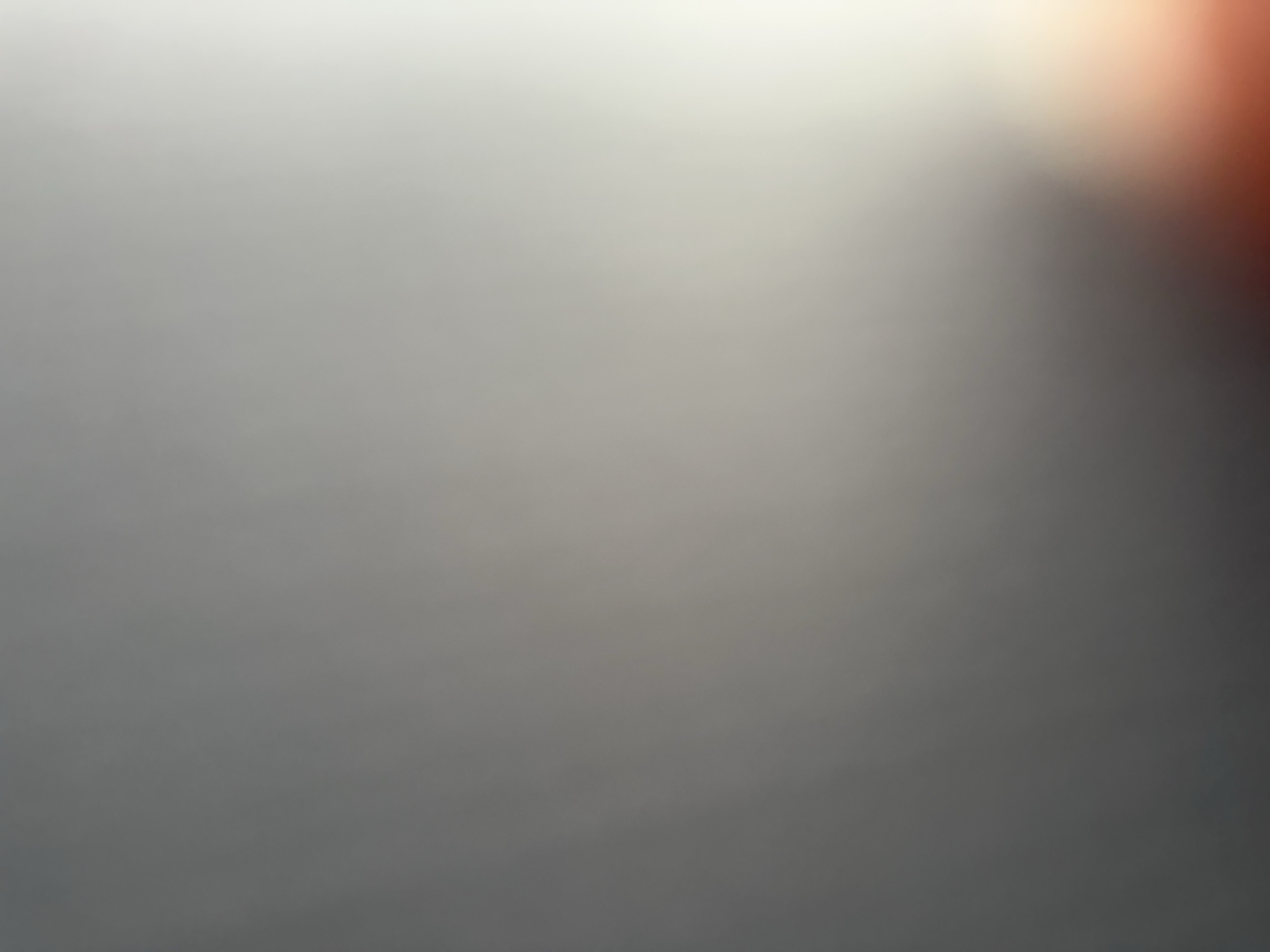 The image is a close-up, abstract shot with a soft focus. The dominant colors are various shades of grey, transitioning from light grey at the top to a darker grey at the bottom. In the upper right corner, there is a warm, diffused light source, appearing as a soft orange or red glow that gradually fades into the grey. The lighting creates a gentle gradient across the image, with the top and right sides being brighter and the bottom and left sides darker. There is no discernible subject matter such as people, objects, or a specific scene. The abstract nature of the image makes it difficult to infer any activity or event. There are no visible text elements. Given the lack of specific visual cues, it is not possible to determine the time of day, weather, or specific location, other than the provided context of Belgrade, Serbia. The overall impression is one of soft light and subtle color variation, creating a calming or atmospheric effect.
