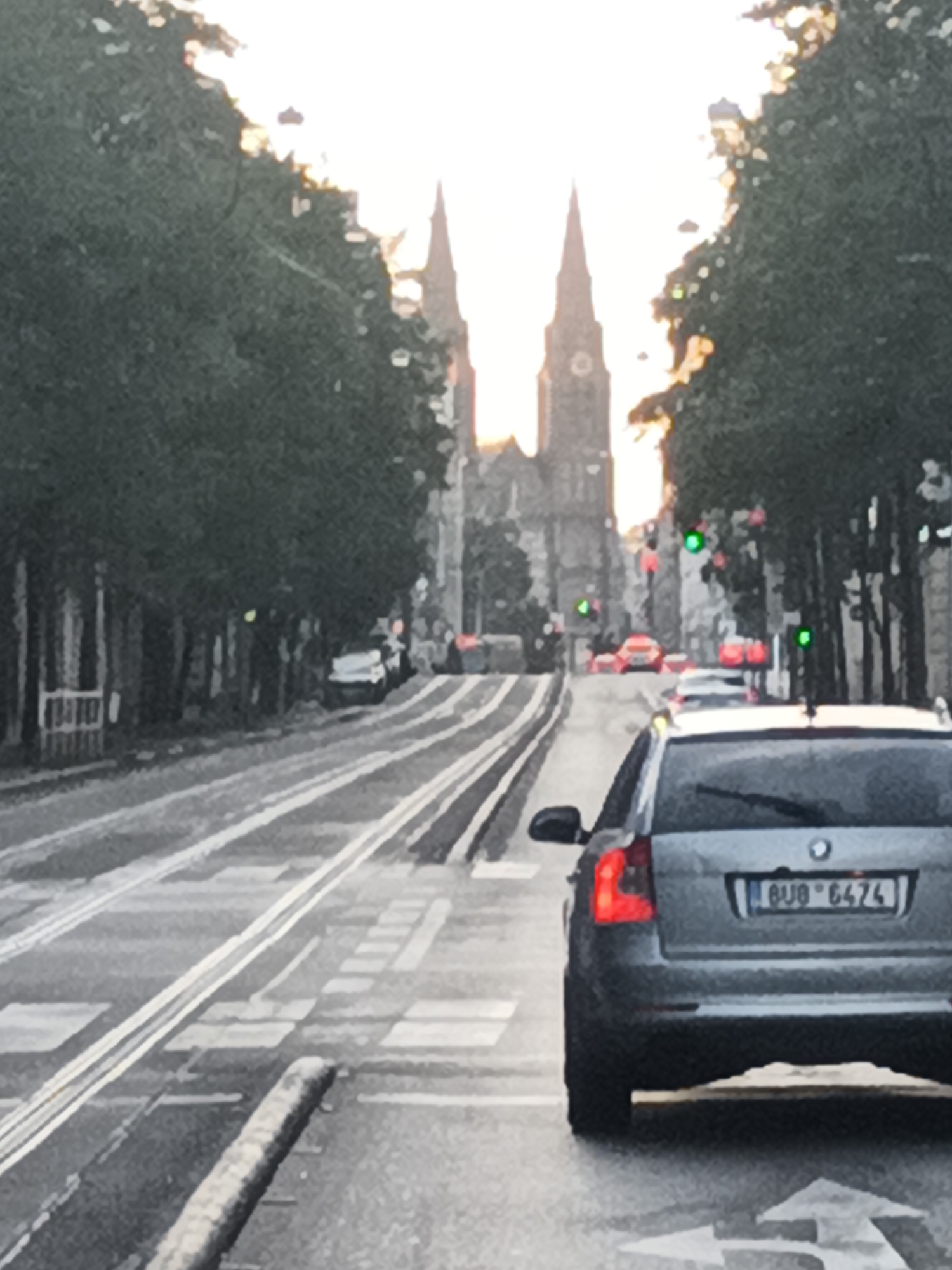 The image depicts a street scene in Prague, Czechia, focusing on a gray BMW SUV from behind, positioned at a pedestrian crossing. The car's license plate reads "6U0 6474". The street features tram tracks, and traffic lights are visible in the background, some displaying green. Several other vehicles are present further down the road, along with trees lining the street. In the distance, two prominent church spires rise above the buildings, characteristic of Prague's architecture, hinting at a location near a significant religious or historical site, possibly St. Vitus Cathedral or another notable church within the city. The sky appears bright, suggesting it is either sunrise or sunset.
