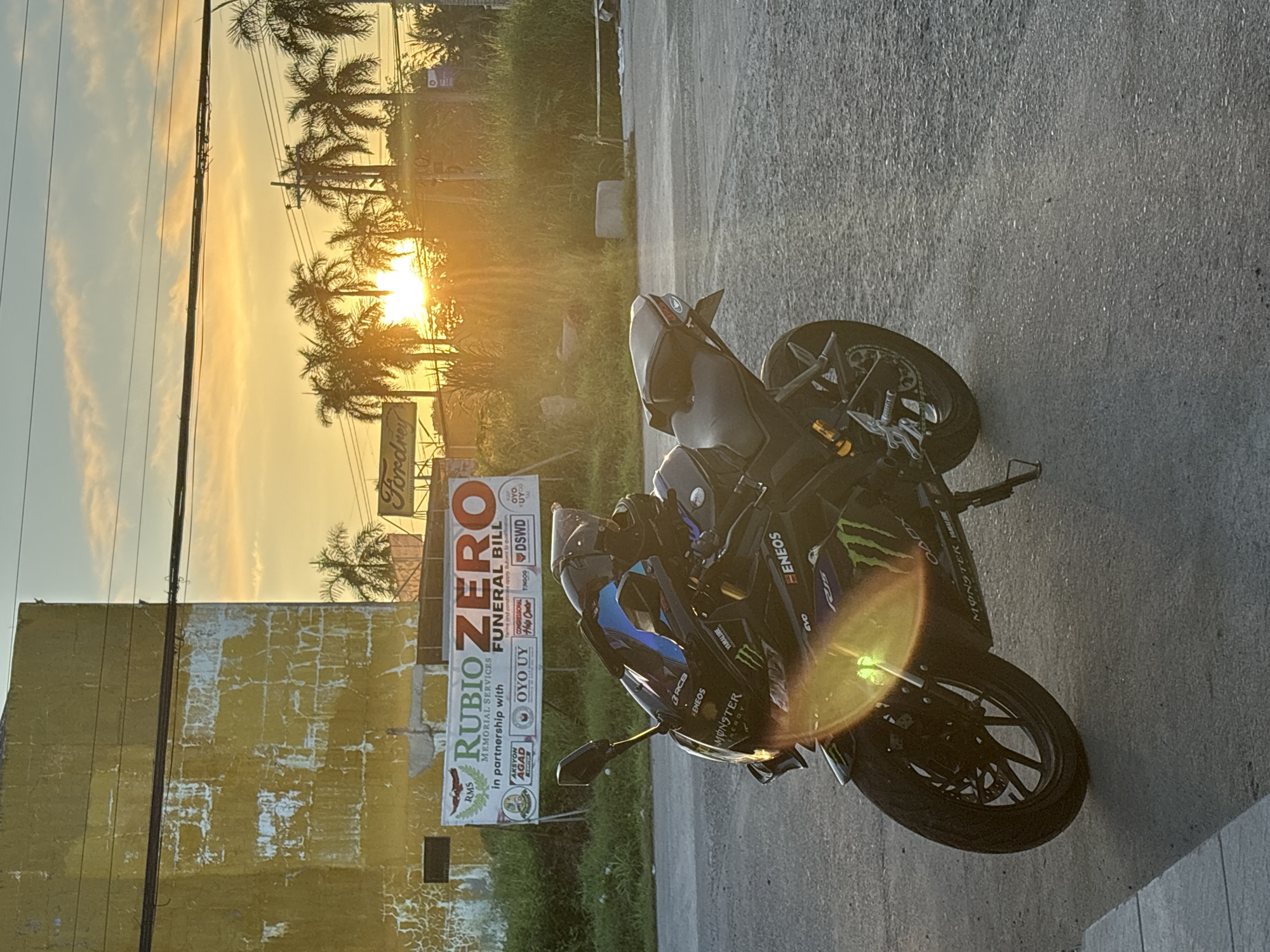 An eye-level, medium shot captures a black Yamaha R15 motorcycle parked on an asphalt road at dusk in Tagum City, Philippines. The motorcycle, adorned with Monster Energy decals and other sponsor logos such as Eneos and RCB, faces the right of the frame. The setting sun, a bright orange orb, peeks through a cluster of palm trees in the background, casting a warm glow and lens flare across the scene. Behind the motorcycle, a weathered yellow building with peeling paint stands to the left. A banner is prominently displayed on the building, advertising "RUBIO ZERO FUNERAL BILL" in partnership with various entities, including RMS, Aksyon Agad, OYO UY, Congressional Help Center, Tingog, and DSWD. Power lines are visible across the top of the frame, contributing to the urban landscape. The ground is a mix of asphalt and gravel, with a concrete sidewalk visible in the lower left corner. The overall atmosphere is serene, illuminated by the fading sunlight.