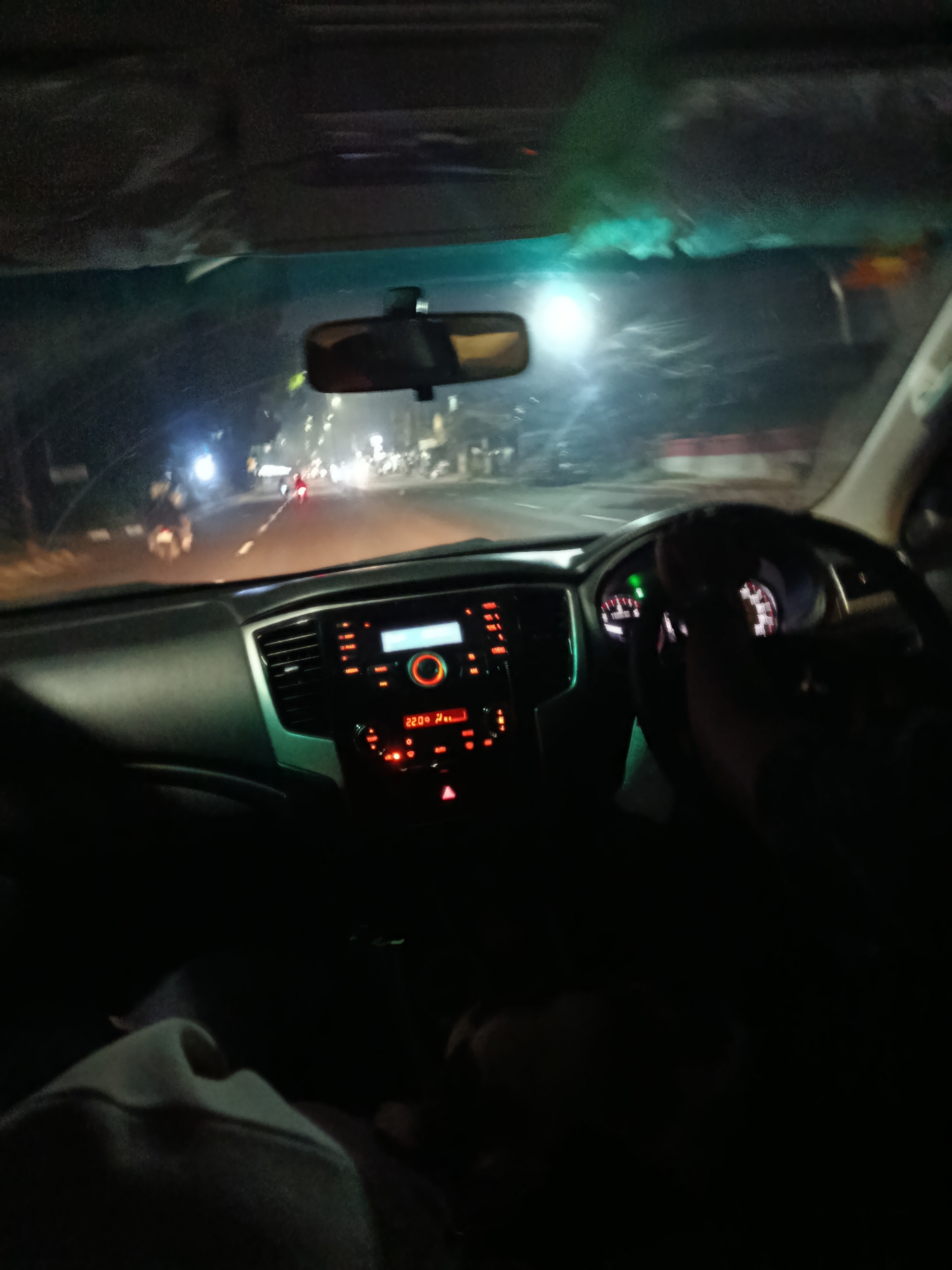 The image depicts the interior of a car driving at night on a road in Cibinong, Indonesia. The dashboard is visible, with illuminated controls and a digital display showing "22.0°". The steering wheel is in the foreground, and a hand is on it, indicating the driver. Outside the car, streetlights and the headlights of other vehicles are visible, suggesting an urban environment. The windshield shows the street and traffic ahead. The scene appears to be captured from the passenger seat.
