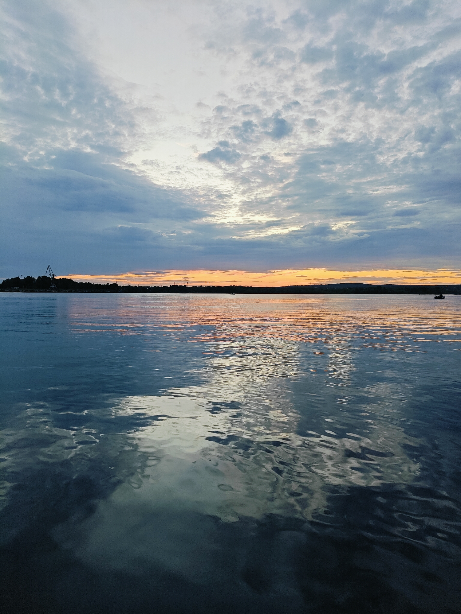 The image depicts a sunset over the Kama River in Chaikovsky, Russia. The sky is filled with a mix of gray and white clouds, with a bright orange band of light visible at the horizon, reflecting on the water. The river surface is calm with gentle ripples, and the reflections of the sky and clouds are visible. On the left side of the horizon, a silhouetted industrial crane stands against the tree-lined shore. In the distance, there are a few small boats on the water.