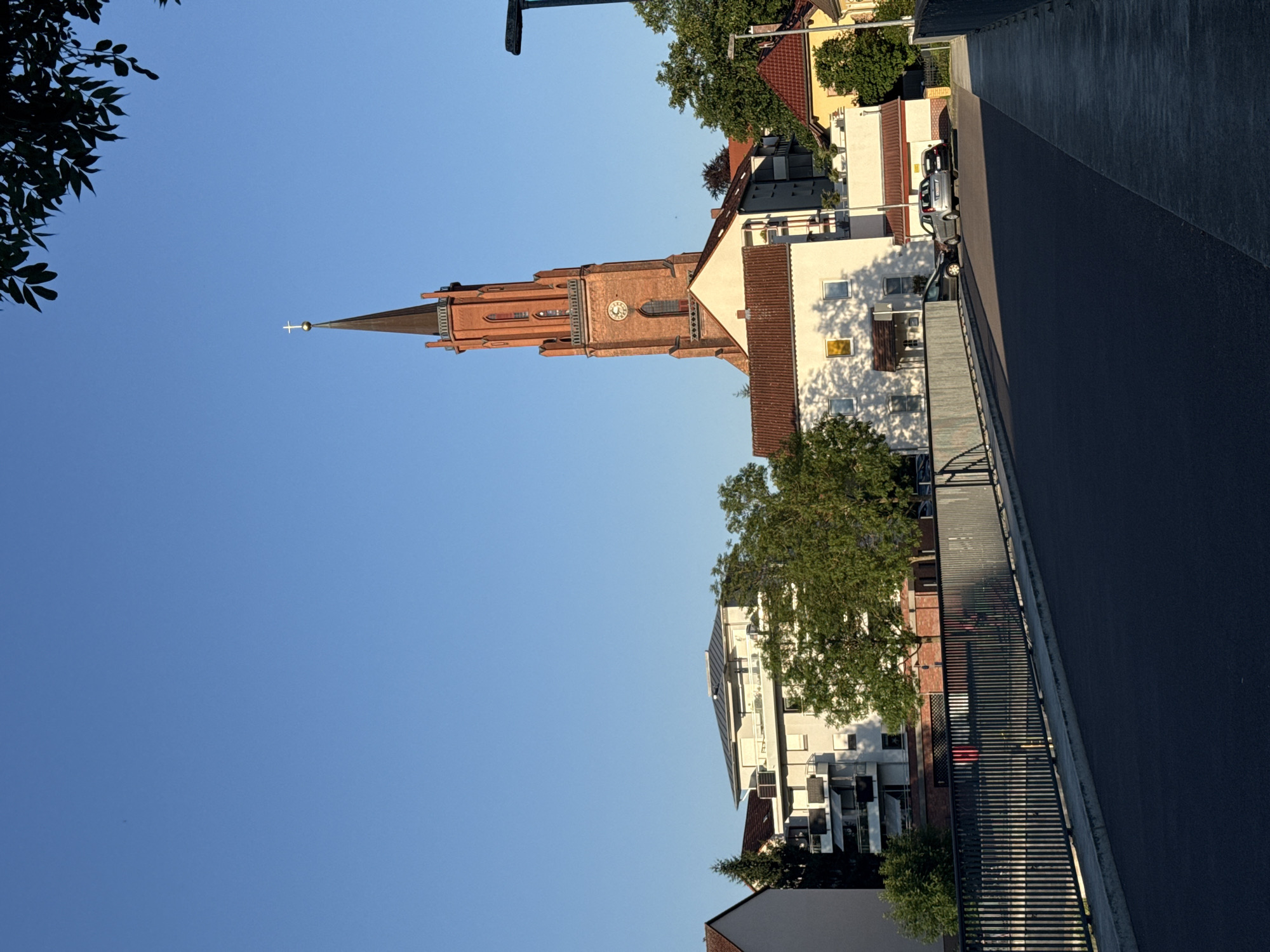 An outdoor, full, eye-level, wide shot captures a clear, blue sky above a European-style town with a prominent red brick church tower. The tower is on the right side of the frame and features a clock face and a cross at its peak. In the middle ground, there are several buildings, including a modern white apartment building with balconies, and smaller houses with red-tiled roofs. Trees with green foliage are interspersed among the buildings. In the foreground, a paved road runs across a bridge with a metal railing. Two cars are parked on the road. The lighting suggests it is late afternoon or early morning, with long shadows cast across the scene.