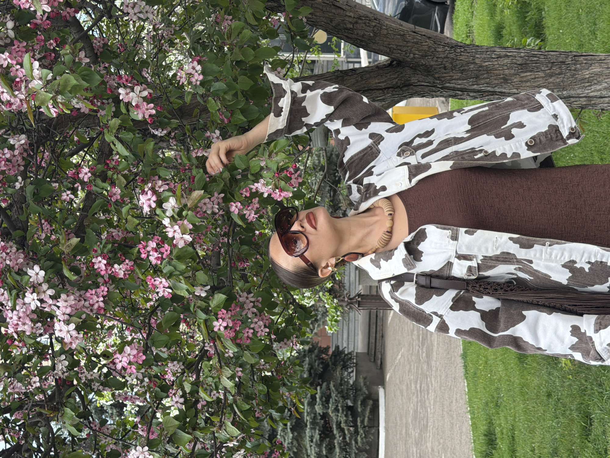 A woman wearing a brown dress, a brown and white cow print jacket, sunglasses, and a necklace stands near a tree with pink flowers. Her right hand is extended towards the flowers. A tree trunk is visible behind her to the right. A sidewalk and grass are visible in the background. The city is Krasnoyarsk, Russia.