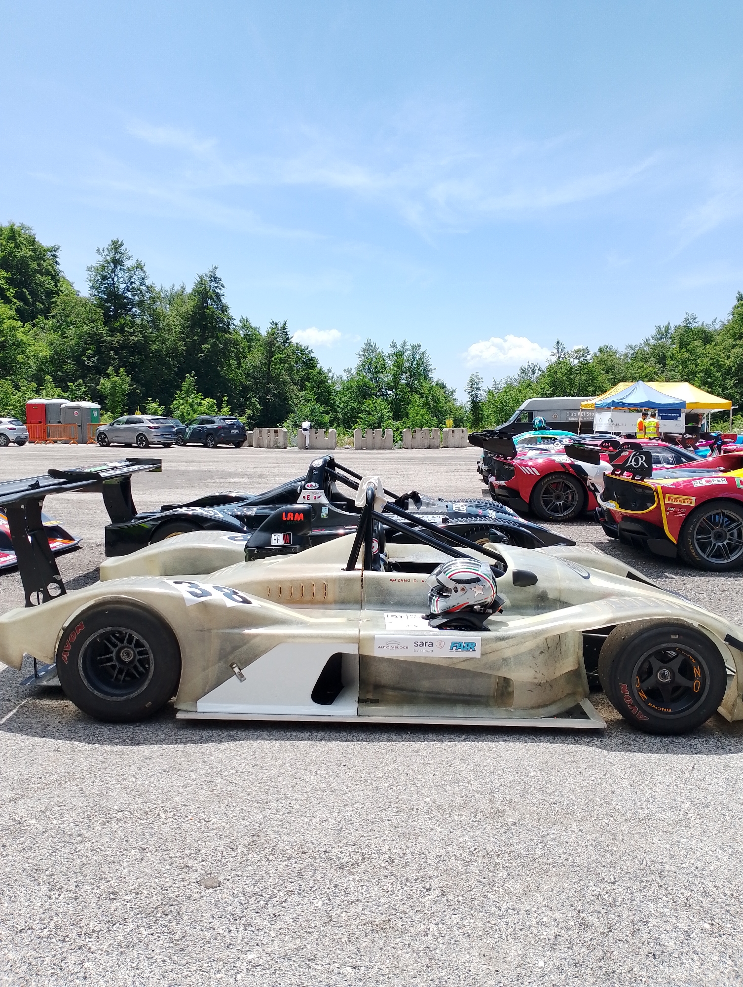 The image depicts a row of open-wheel race cars parked on an asphalt surface, likely a racetrack or pit lane, under a bright, partly cloudy sky. In the foreground, a light-colored, unfinished race car with the number "38" is prominent. A racing helmet rests on its cockpit. Behind it, several other race cars are visible, including a black car with "LAM" and "BELVA" logos, and a red and yellow car with "REOFER" and "PIRELLI" branding. In the background, a yellow and blue tent is set up, with several people milling about. Beyond the track, a dense line of green trees borders the area. The presence of portable toilets and barrier walls suggests this is a temporary setup, common for racing events. The overall atmosphere suggests a racing event is either about to begin or has just concluded. The context provided about Rieti, Italy, places this scene in an outdoor, possibly rural or semi-urban setting within the country, characteristic of motorsport venues.