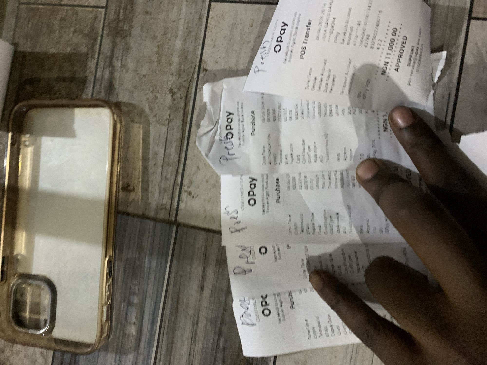 An overhead indoor shot captured in Kabong, Nigeria, displays a dark-skinned human hand interacting with multiple OPay transaction receipts spread across a textured surface. In the upper left quadrant, an empty gold-tinted transparent phone case with a prominent camera cutout is present. The receipts, fanned out in the lower and central portions of the frame, include details of "Purchase" and "POS Transfer" transactions, with one receipt explicitly stating "NGN 11,000.00 APPROVED". Several receipts bear handwritten annotations, possibly "Presh," at their tops. The hand's fingers are positioned directly over the receipts, suggesting examination or organization. The background surface exhibits a brownish-grey, wood-grain or tiled pattern.