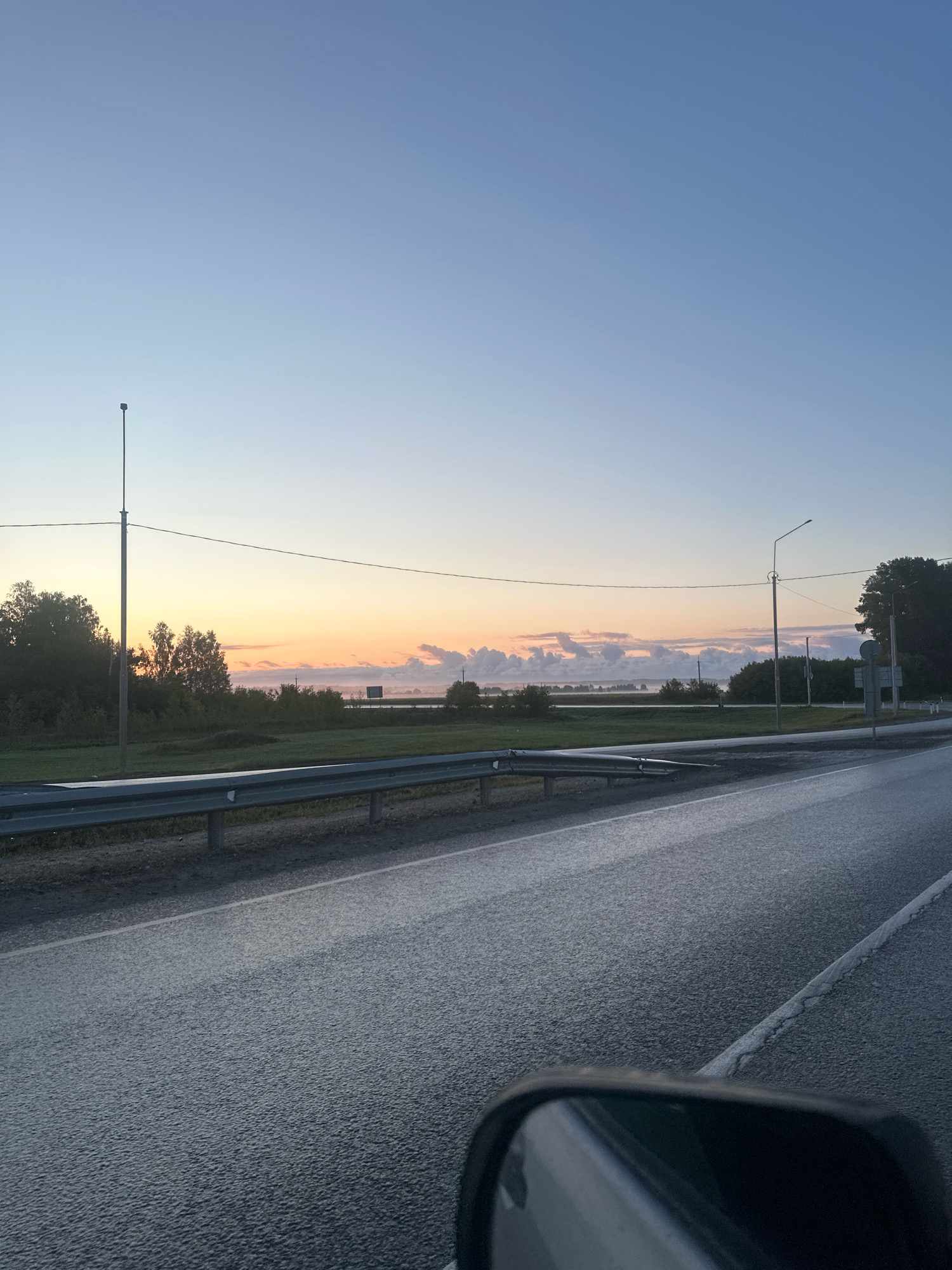 This is a photograph taken from a vehicle, likely while it is in motion on a paved road. The perspective is from the driver's or passenger's side, as a portion of a side mirror is visible in the lower right corner. The road ahead is wet, reflecting the muted light of dawn or dusk. A guardrail runs along the left side of the road, separating it from a grassy verge. Beyond the verge, a line of trees forms a dark silhouette against a sky that transitions from pale blue at the top to soft oranges and yellows near the horizon. Several utility poles with overhead wires are visible, one prominent pole on the left and another further down the road on the right. In the distance, a body of water is visible, with fog or mist clinging to its surface. The sky is mostly clear, with a few scattered clouds near the horizon. The overall impression is one of a tranquil rural or suburban landscape at the beginning or end of the day.