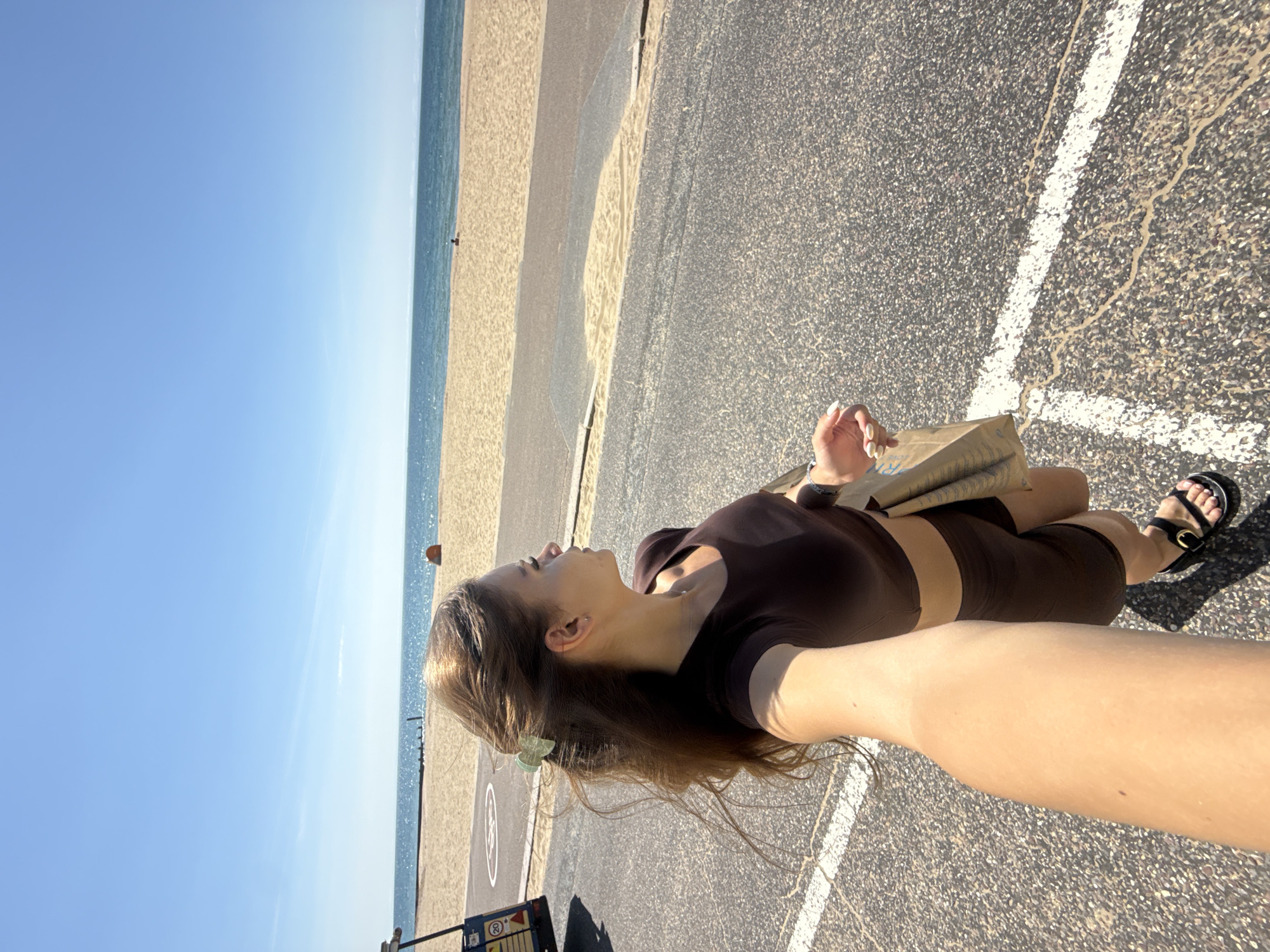 A woman is walking on a paved path beside a beach. The sky is clear blue, and the sea is visible in the background. The woman is wearing a brown crop top and matching shorts, and she has a shopping bag in her hand. She is also wearing black sandals. There is a vehicle on the left side of the image, and some beach huts are visible in the distance on the beach. The scene appears to be a sunny day in Bournemouth, United Kingdom.