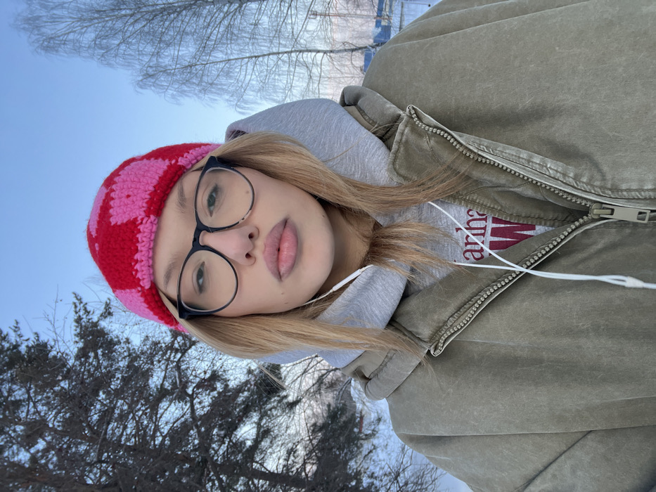 A casual selfie captures a person outdoors on what appears to be a bright winter or early spring day. The image is rotated 90 degrees to the right.
The central subject is a young person, likely female, with long, straight blonde hair framing their face. They are wearing a distinctive red and pink checkerboard or plaid patterned knitted hat/beanie. Large, round-framed black eyeglasses are perched on their nose, reflecting some light. Their lips are slightly parted, revealing a natural pink hue.
The person is dressed in warm, layered clothing. Over a light-colored top, they wear a light grey hooded sweatshirt, with the hood visible around their neck. The outermost layer is a sturdy-looking, muted olive green or khaki-colored jacket with a prominent central zipper, which is partially open. White wired earphones are visible, with one earbud in the left ear and the wire trailing down across their chest. Beneath the grey hoodie, a portion of a white t-shirt is visible with the brand name "Carhartt" printed in a dark red or maroon font.
The background consists of a clear, bright blue sky, suggesting fair weather. Bare tree branches, some appearing thin and delicate, are silhouetted against the sky, possibly covered with a light dusting of snow or frost, indicating cold temperatures. Further down, darker, possibly evergreen, trees are visible, adding depth to the scene. The overall impression is one of a relaxed outdoor moment on a cool, clear day.