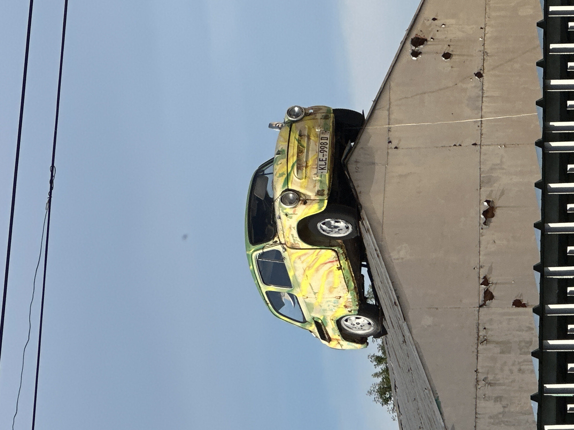 A weathered, yellow and green painted vintage car is perched precariously on the peak of a sloped, gray metal roof of a building. The car, identified by its license plate "KLE-998 D," appears to be a Fiat 600 or a similar model from the mid-20th century. The vehicle exhibits signs of age, including rust and peeling paint, contributing to its artistic or whimsical presentation.
The building beneath the car has a light-colored, possibly concrete or stucco, facade with visible patches of damage and peeling paint. The roof is made of corrugated metal sheets. In the foreground, a dark fence with vertical supports is partially visible.
The background consists of a clear, pale blue sky with two dark power lines running horizontally across the top of the frame. To the left, a small cluster of green trees can be seen behind the roofline. The scene suggests an unconventional display or art installation located in Lyaskovets, Bulgaria, given the context provided. There are no people visible in the image.